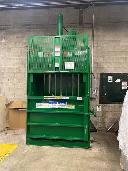 Green industrial cardboard compactor in a concrete-walled room.