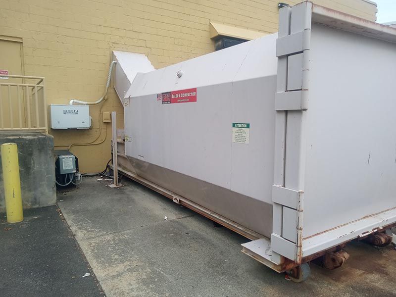 Large industrial waste compactor against a light yellow building, parked on concrete.