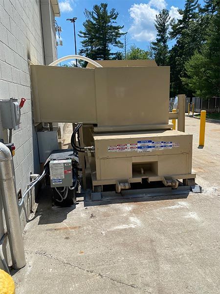 Beige industrial trash compactor next to a building. Electrical components visible, outdoors on concrete.