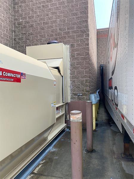 Trash compactor next to a semi-truck against a brick wall; narrow alleyway.