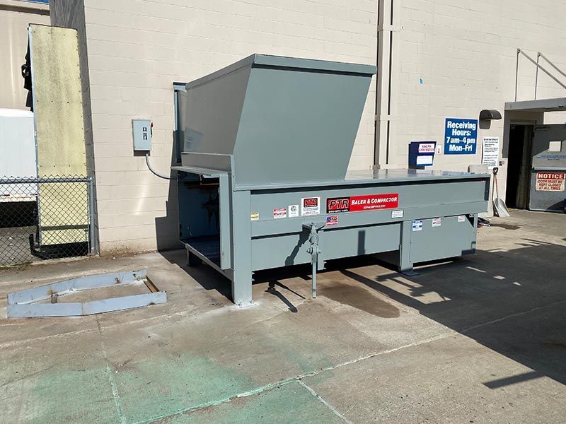 Gray industrial trash compactor outside building, angled view.