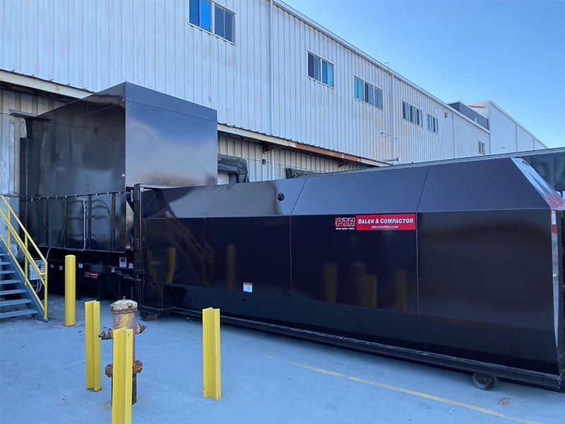 Black industrial compactor outside a building, next to a truck, on a loading dock.