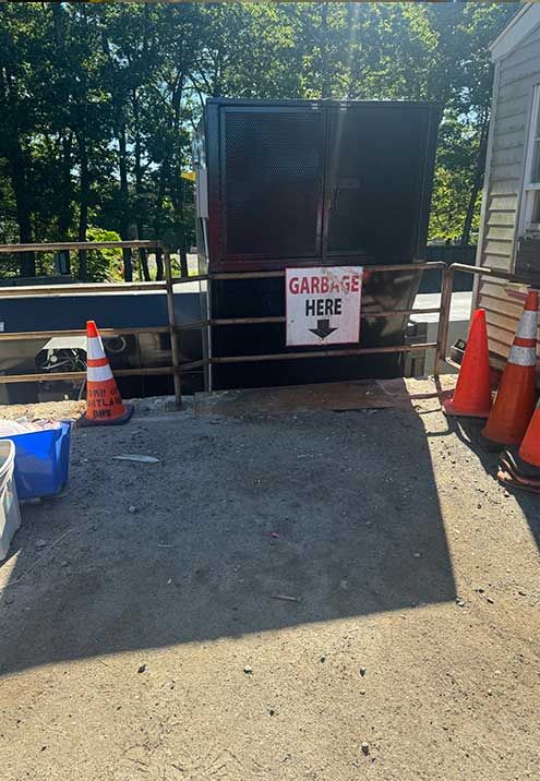 Gare-age sign points to a black dumpster behind a metal fence, set outdoors with orange cones.