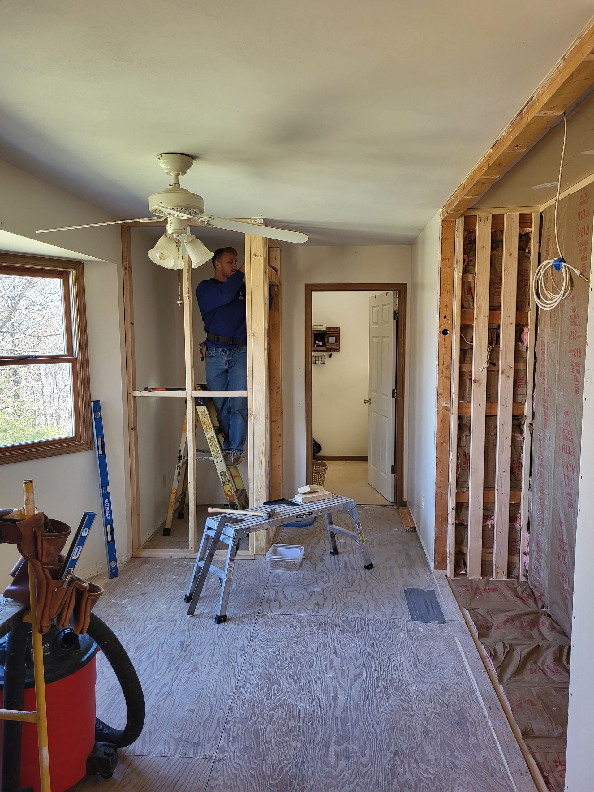 A man is standing on a ladder in a room with a ceiling fan.