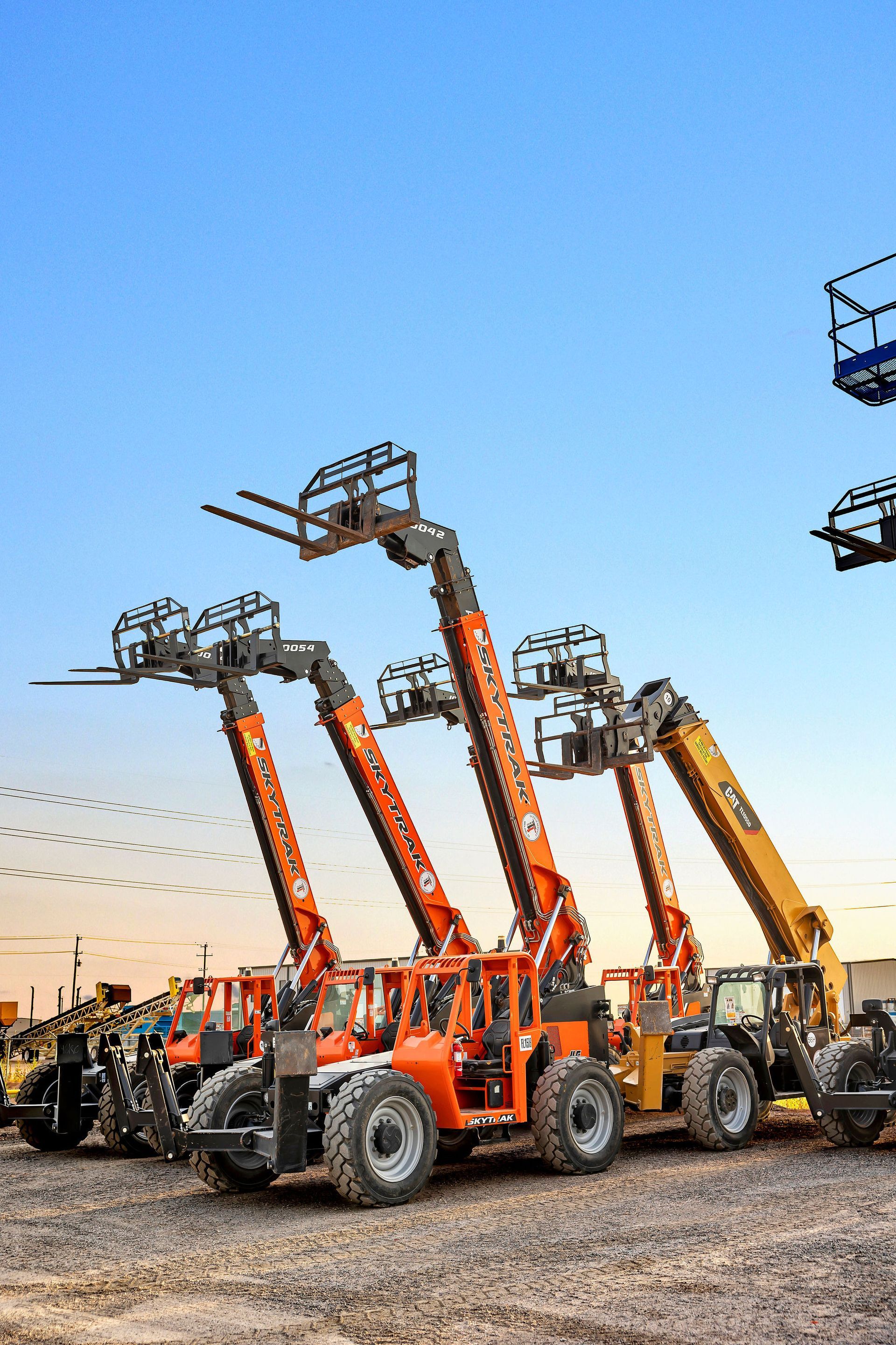 Orange and yellow telehandlers lined up under a blue sky in a construction equipment yard.