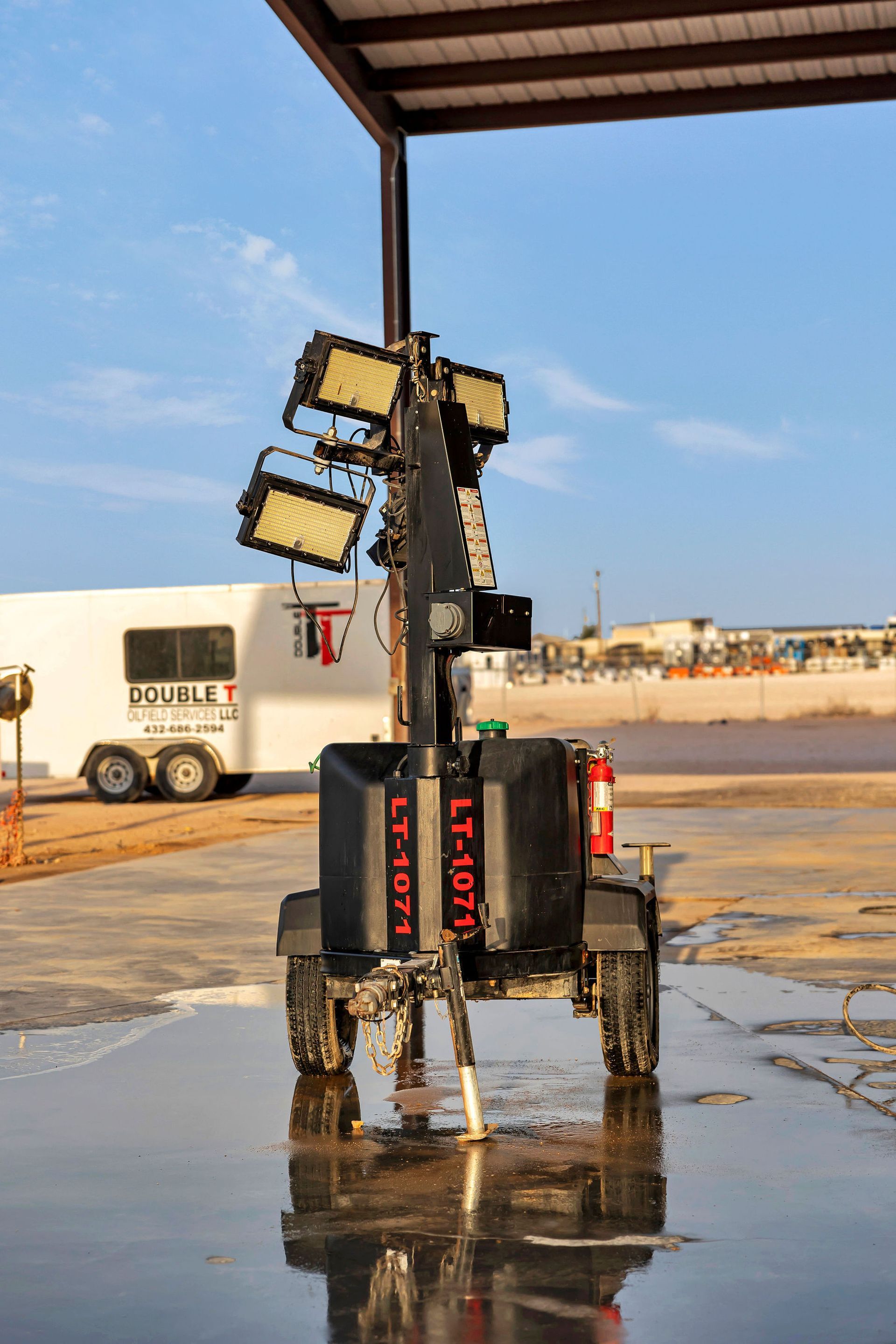 Mobile light tower with bright lights, standing under a canopy on a wet surface; trailer in the background.