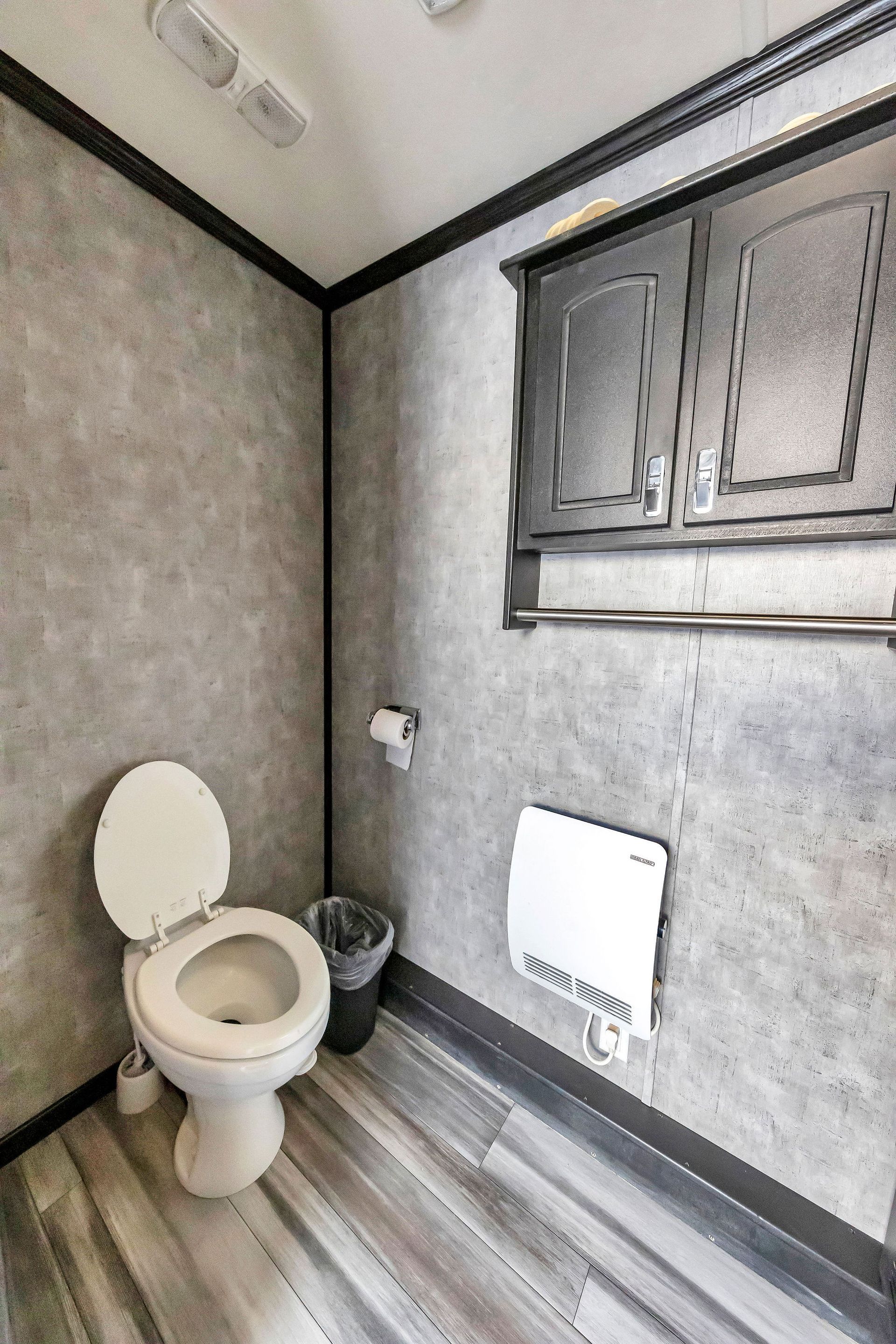 A small, gray-walled bathroom with a toilet, cabinet, and floor heater; toilet paper roll on wall.