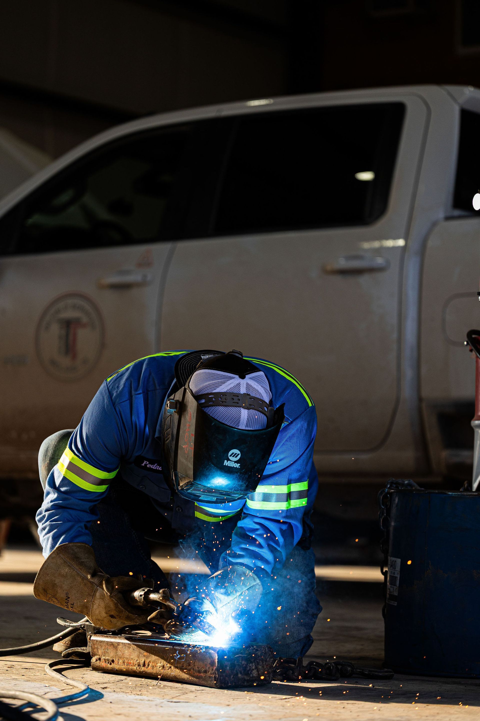 Welder in blue coveralls, mask on, working on metal with bright blue sparks, truck in background.
