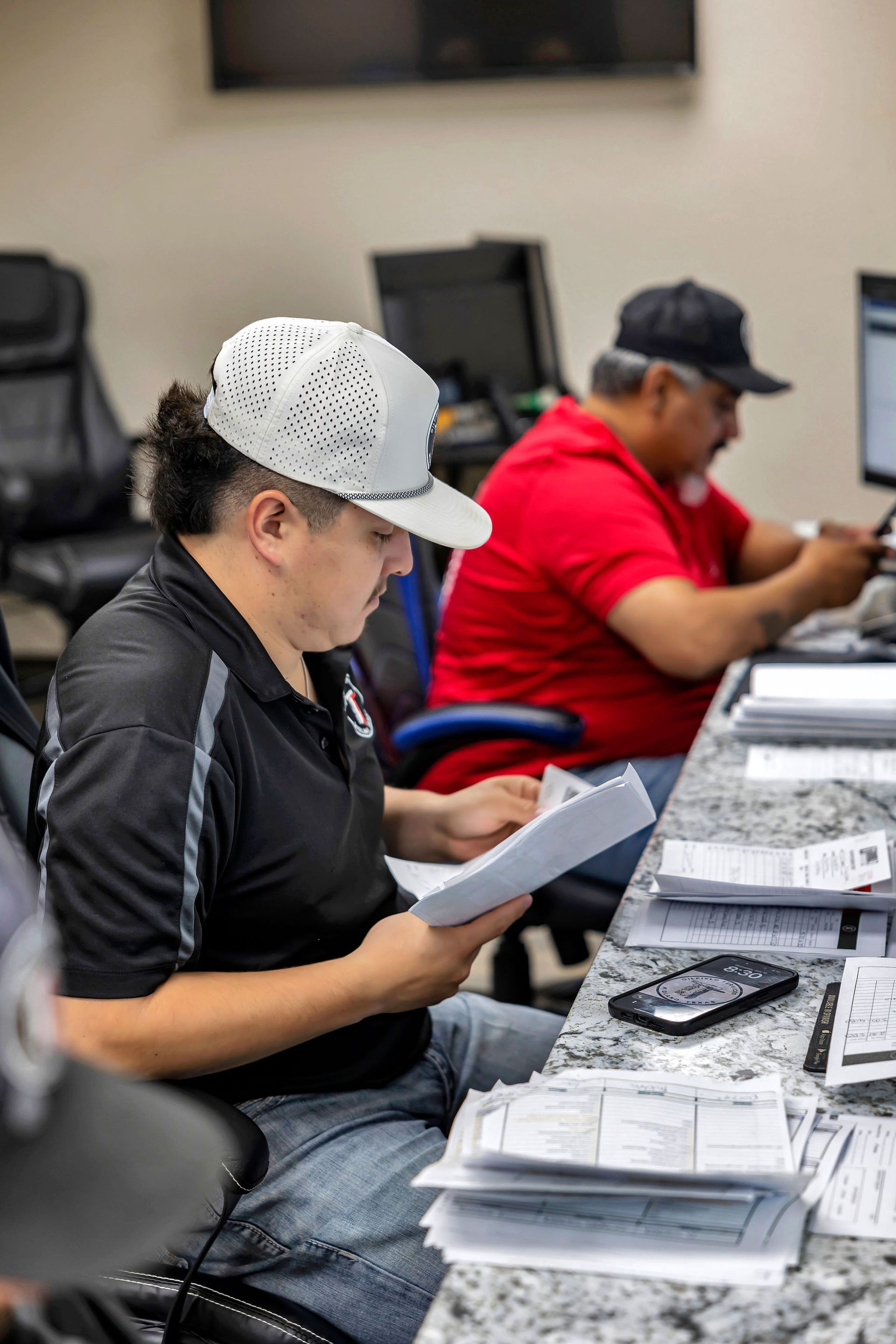 Two men reviewing documents at a table. One wears a white hat, the other a red shirt. Office setting.