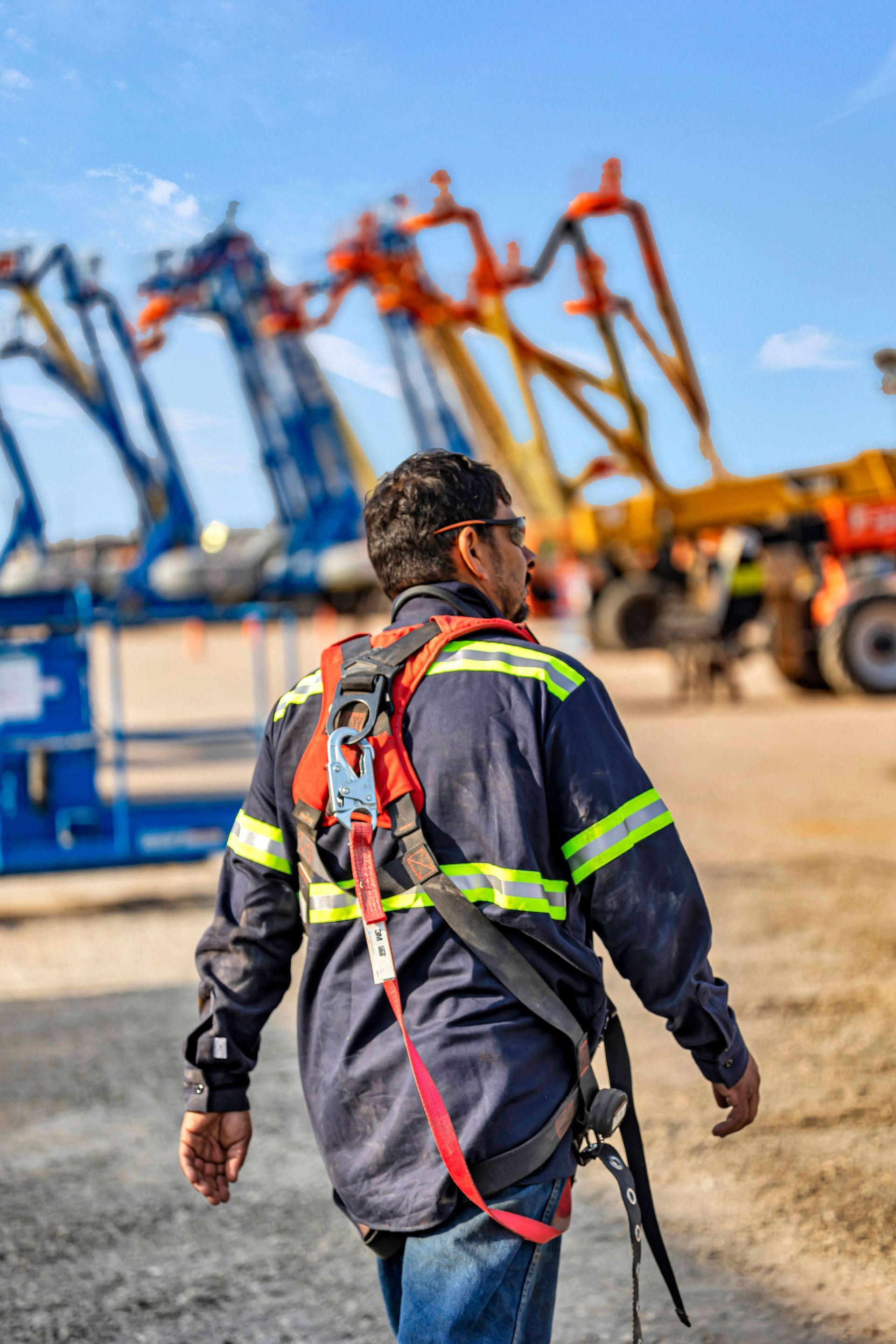 Construction worker in safety harness walking towards industrial machinery.