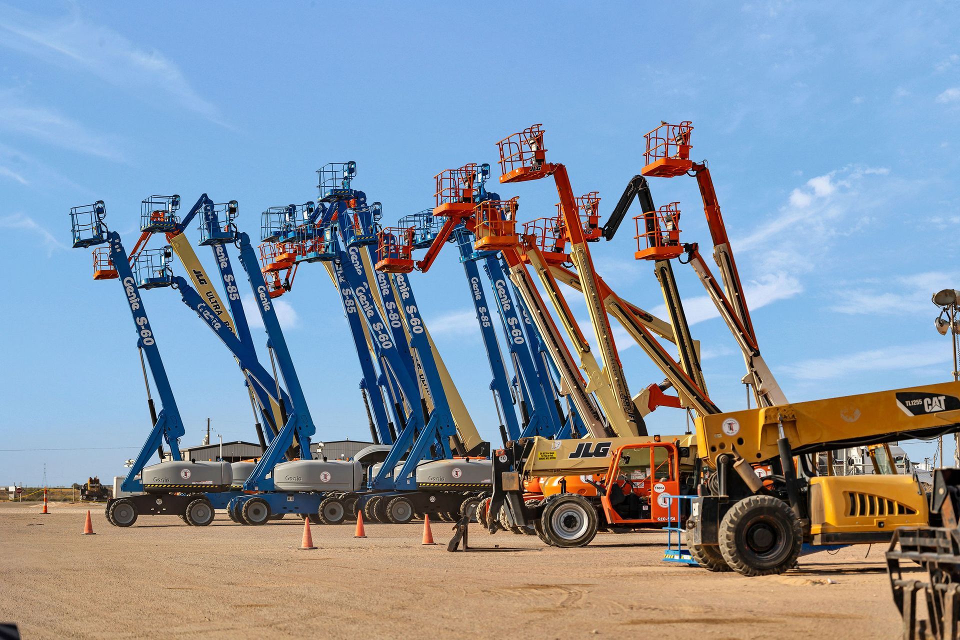 Blue and orange boom lifts parked in a dirt lot under a blue sky.