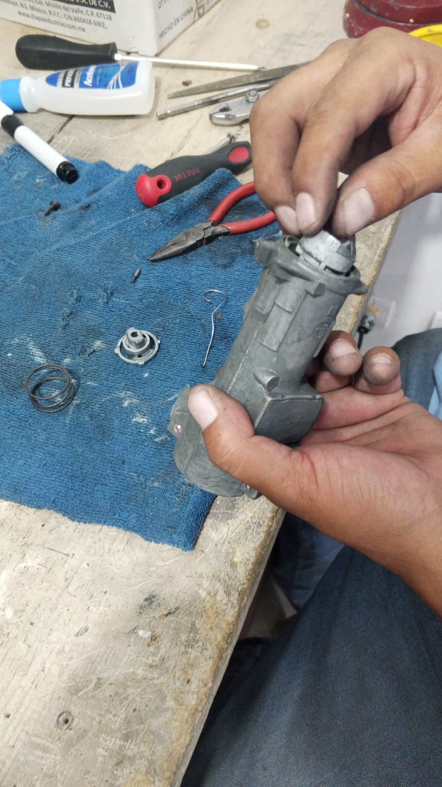 Manos ensamblando un cilindro de cerradura de encendido de un vehículo en un banco de trabajo con herramientas.