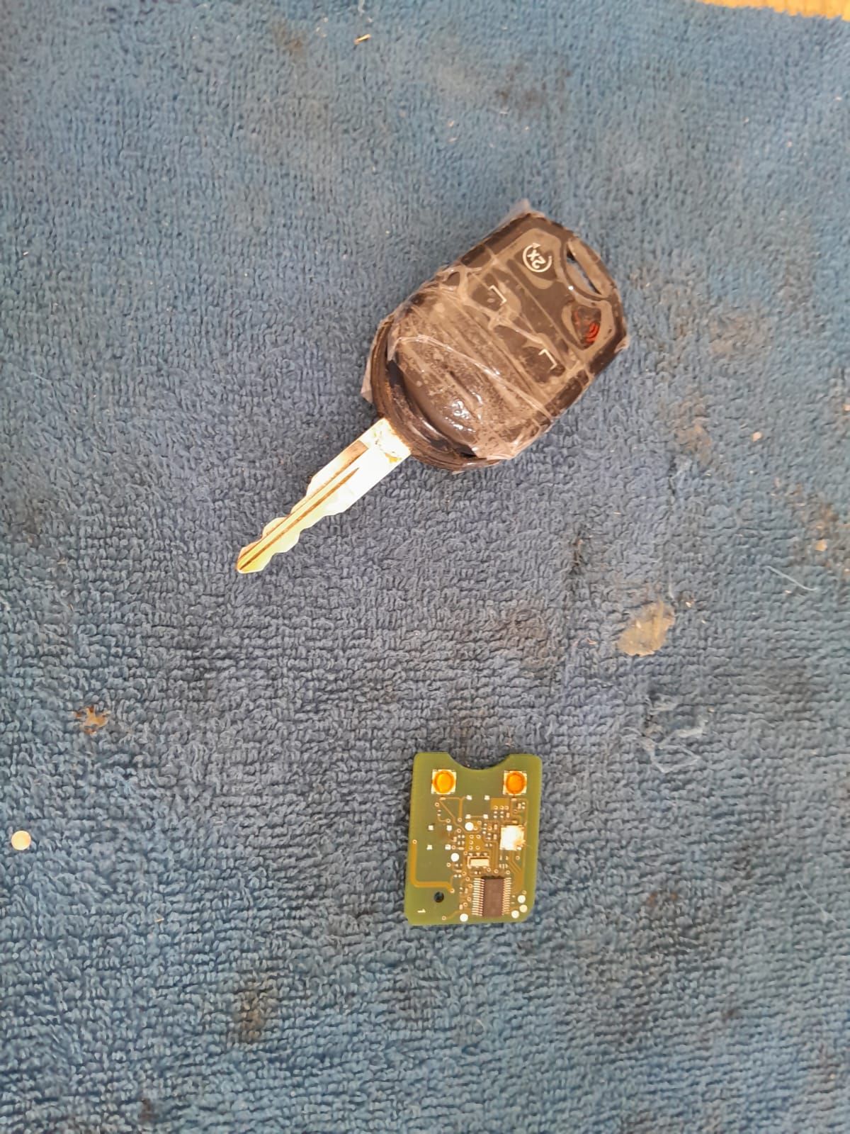 Llave de coche envuelta en cinta negra, con una pequeña placa de circuito, sobre una superficie de tela azul.