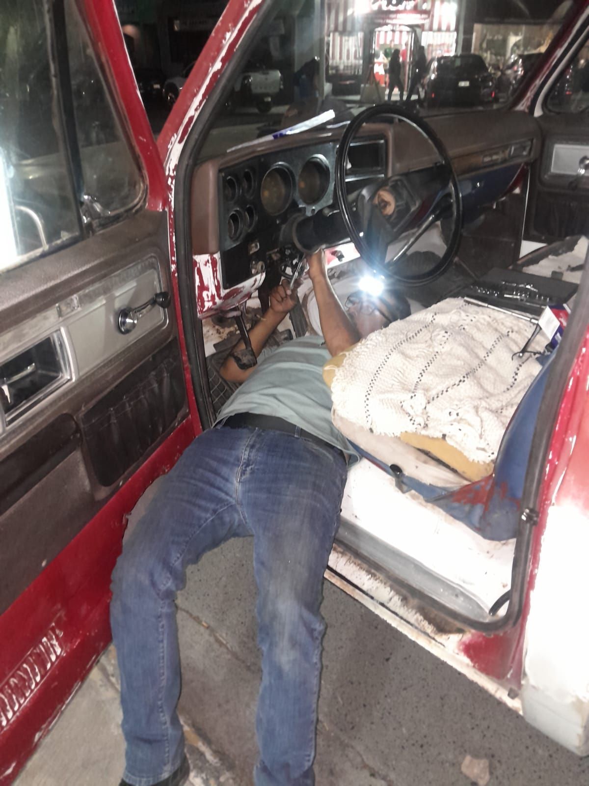 Persona trabajando en el tablero de instrumentos de un automóvil rojo, acostado con una linterna en un garaje.