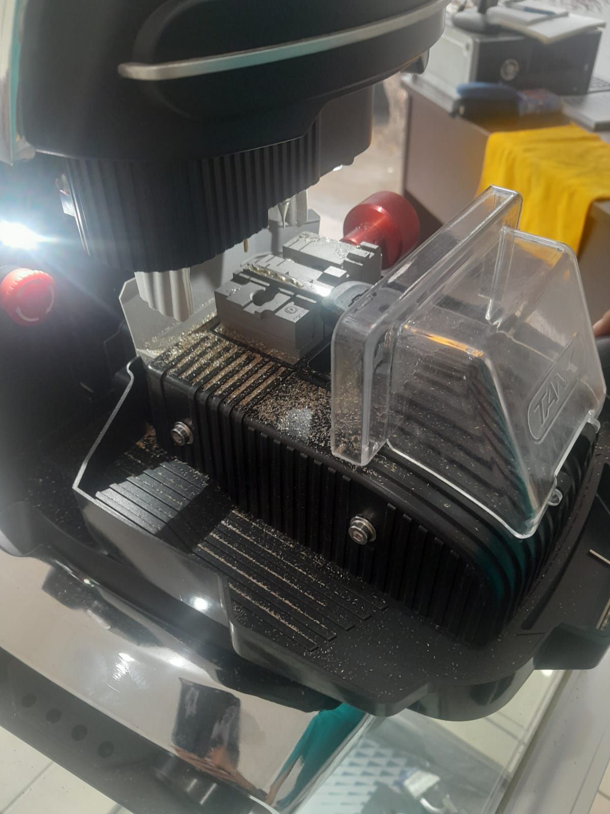 Maquina duplicadora de llaves, de color negro, con guardapolvo, protector de seguridad transparente, llaves y pulsador rojo.