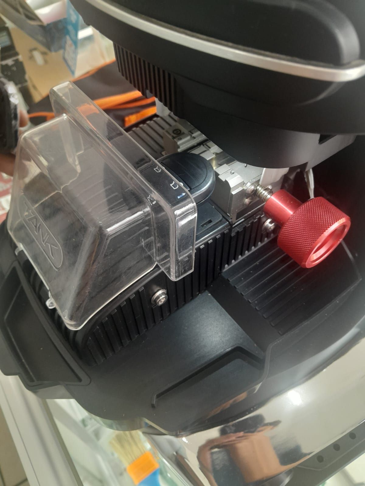 Primer plano de una máquina cortadora de llaves negra con una perilla roja y una cubierta de plástico transparente.