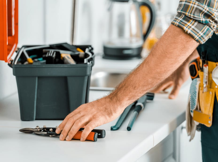 Plumber with tools reaching for pliers on a white countertop. Toolbox and kettle in background.