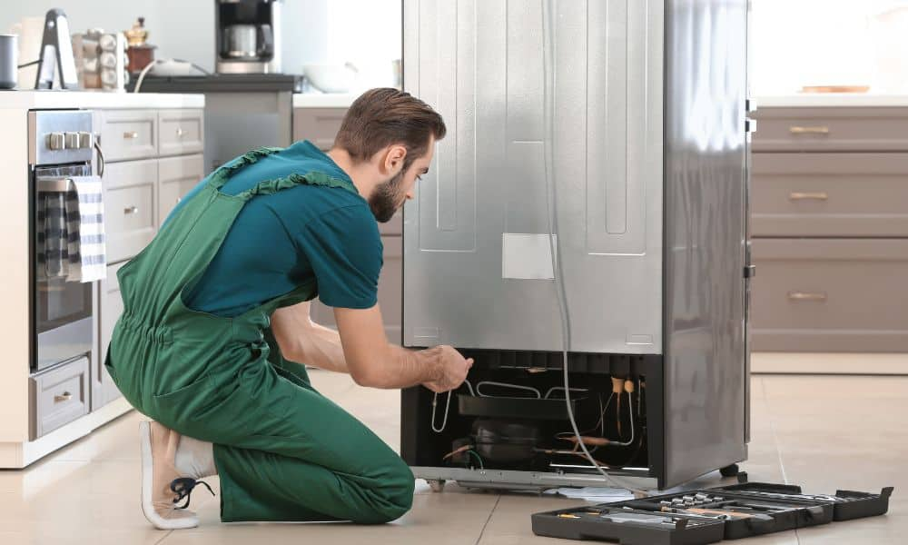A person in green overalls is fixing the back of a refrigerator in a kitchen.