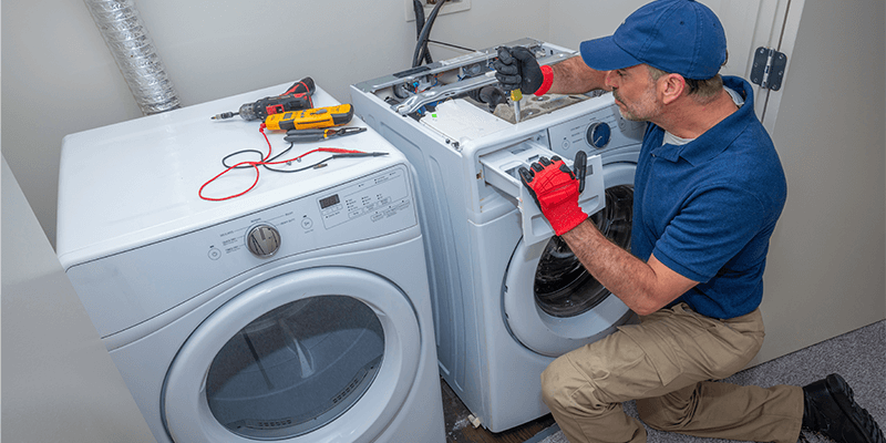 A repairman kneels, working on a washing machine. A screwdriver and tools lie on top of a nearby dryer.