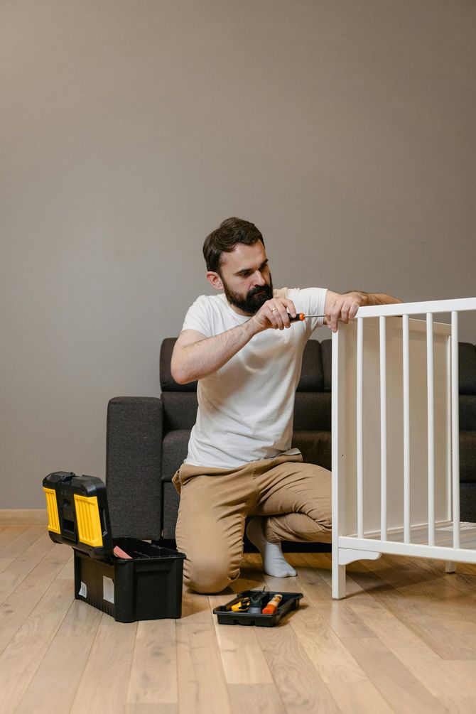Man assembling a white crib with a screwdriver, tools nearby on a wooden floor, gray wall backdrop.