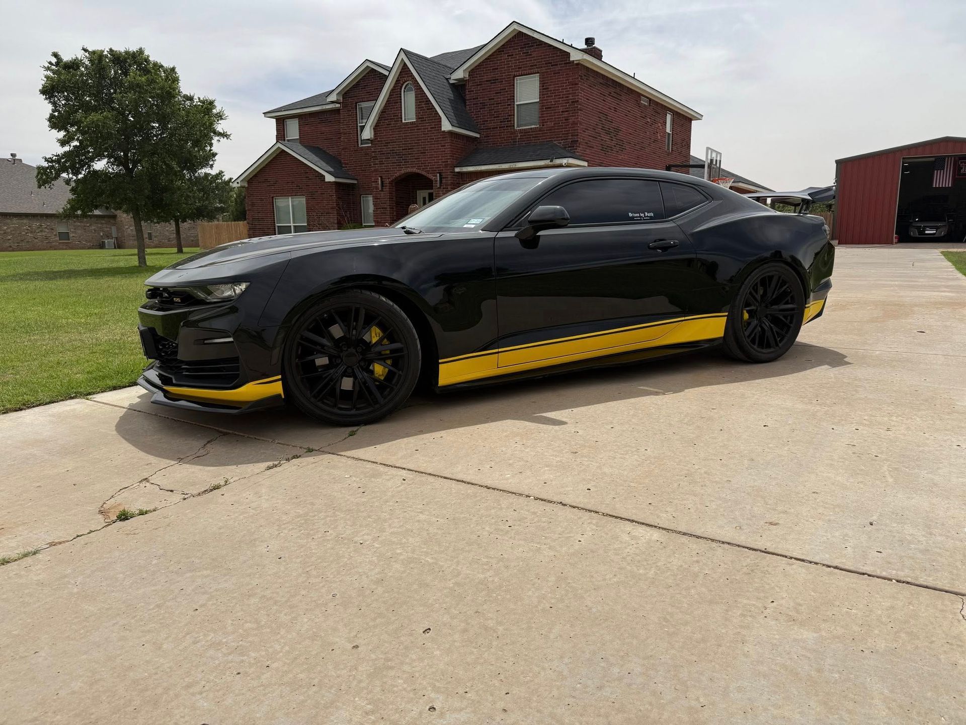 A black and yellow sports car is parked in a driveway in front of a brick house.