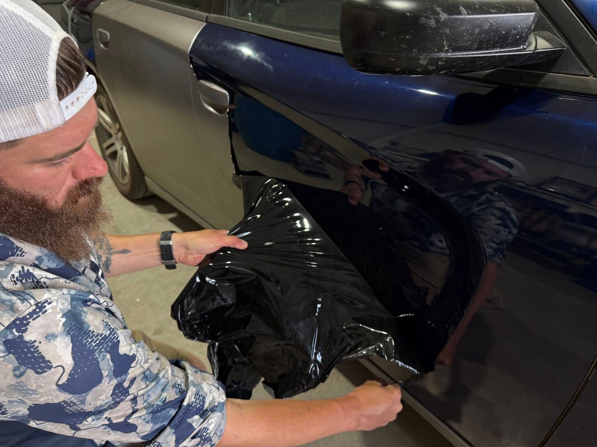 A man with a beard is wrapping a car in black plastic.