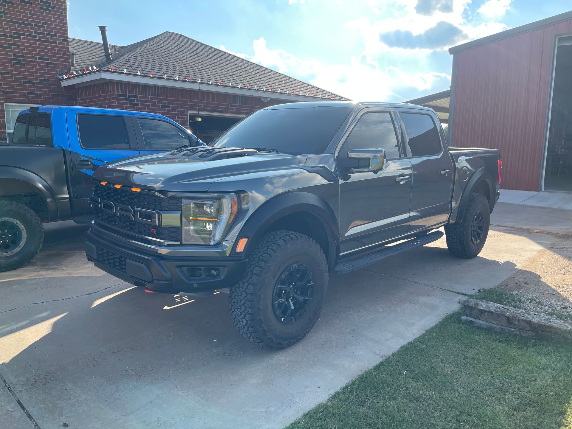 Dark gray Ford Raptor truck parked outside, with a blue truck partially visible behind it. The Raptor has black wheels and tinted windows.