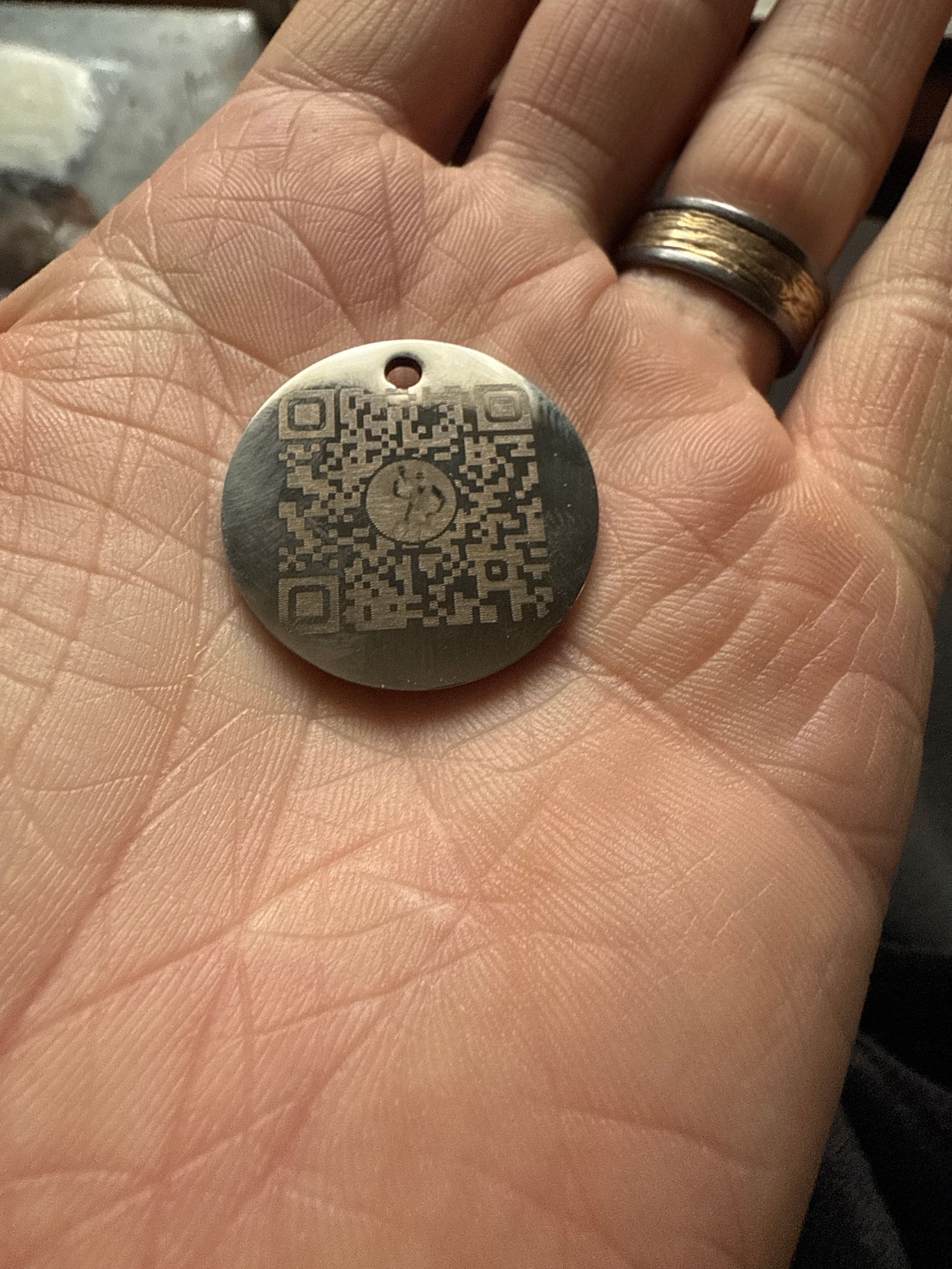 A person's hand holds a circular metal tag with a QR code etched into it. The tag has a small hole for attachment.
