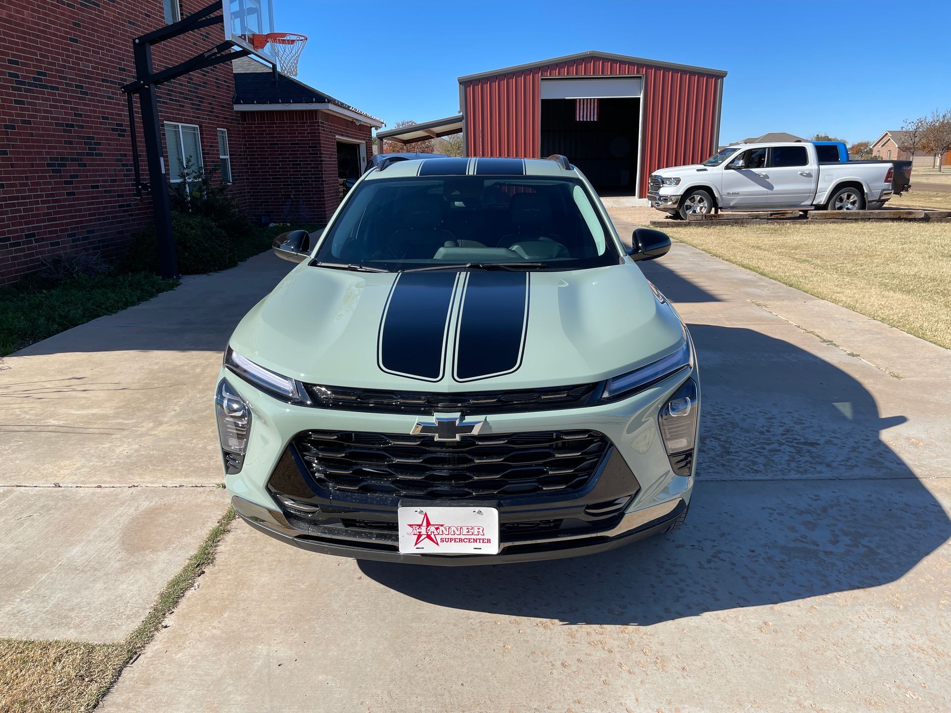 Green Chevrolet Trailblazer SUV with black racing stripes parked in front of a red barn and a brick building on a sunny day.