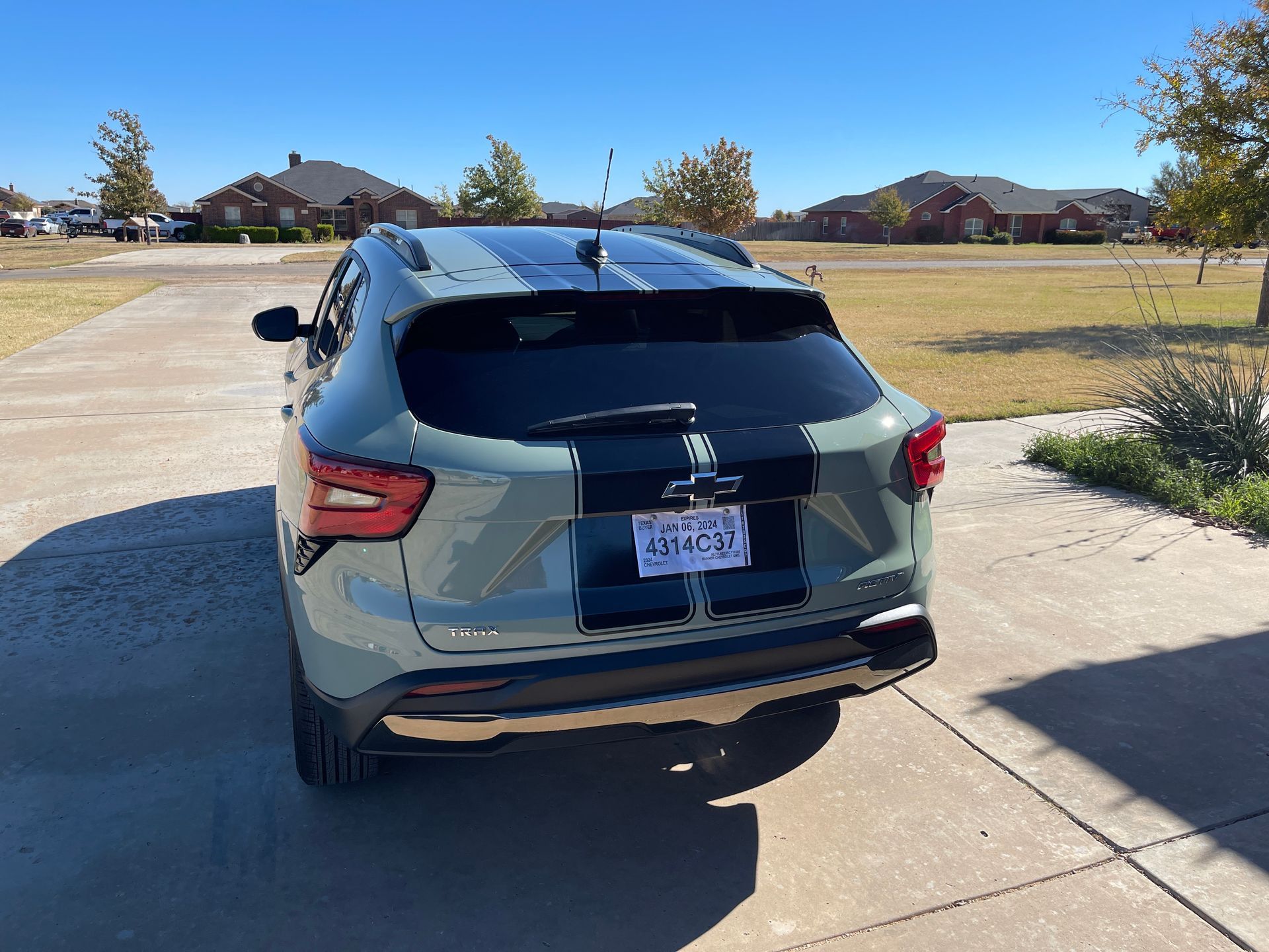 A green SUV parked in a driveway, with black racing stripes, license plate visible. Houses and blue sky in background.
