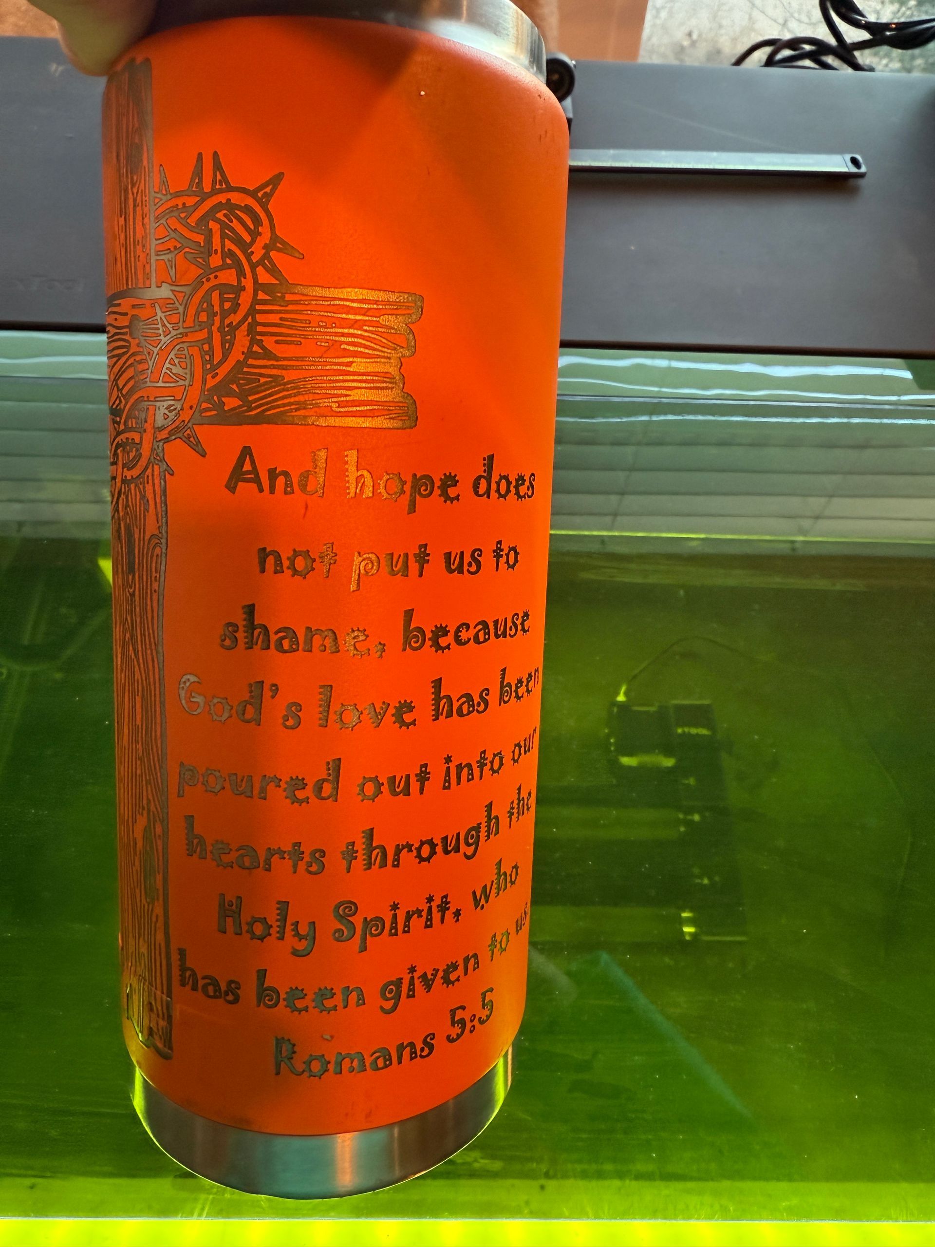An orange tumbler with etched religious text and a cross design. It is held in front of a green-tinted machine.