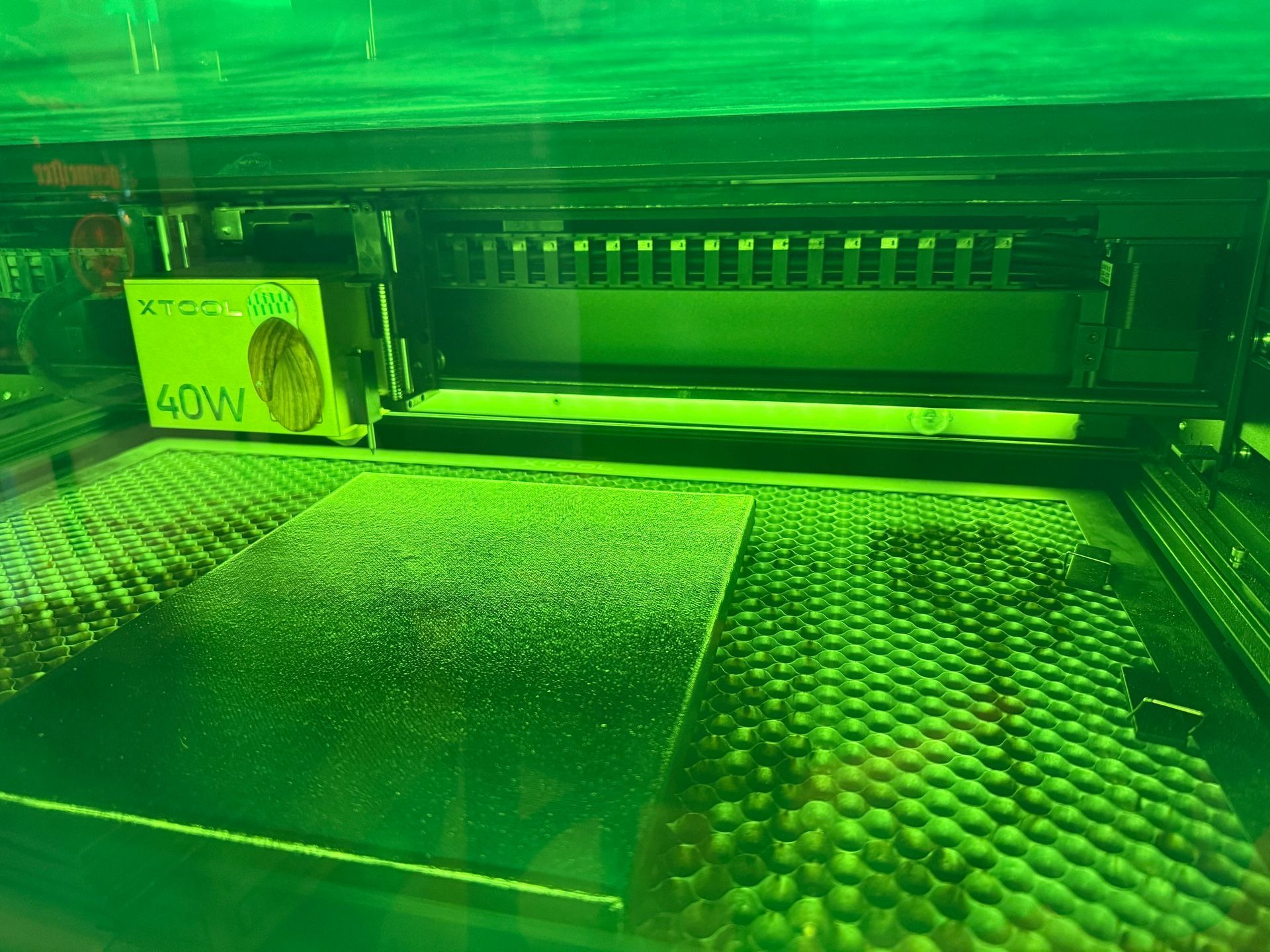 Inside a laser cutter, a green laser illuminates a honeycomb bed. A sheet of material is visible on the bed.
