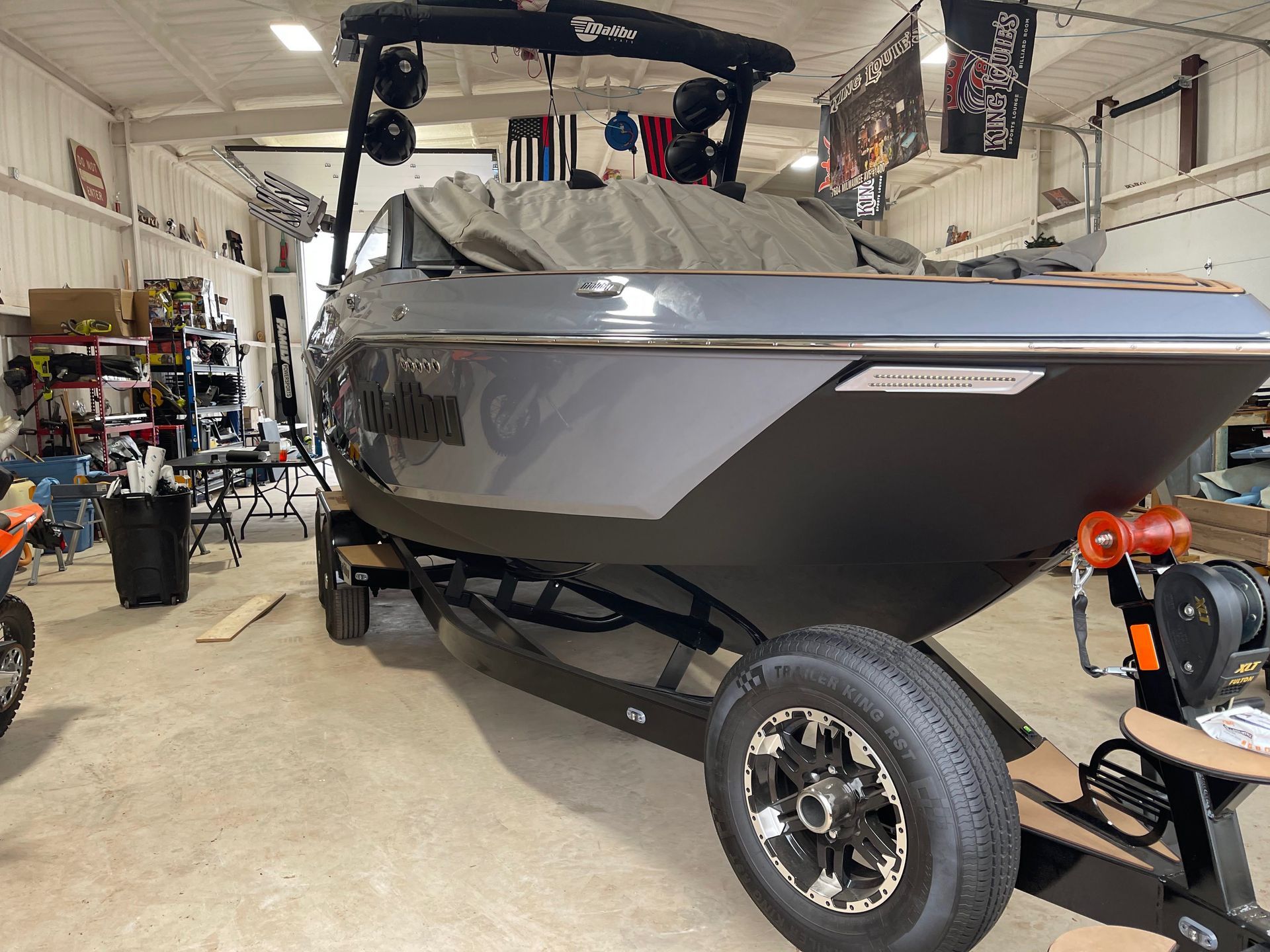 A dark grey wake boat on a black trailer sits inside a garage. The boat is partially covered with a gray tarp.