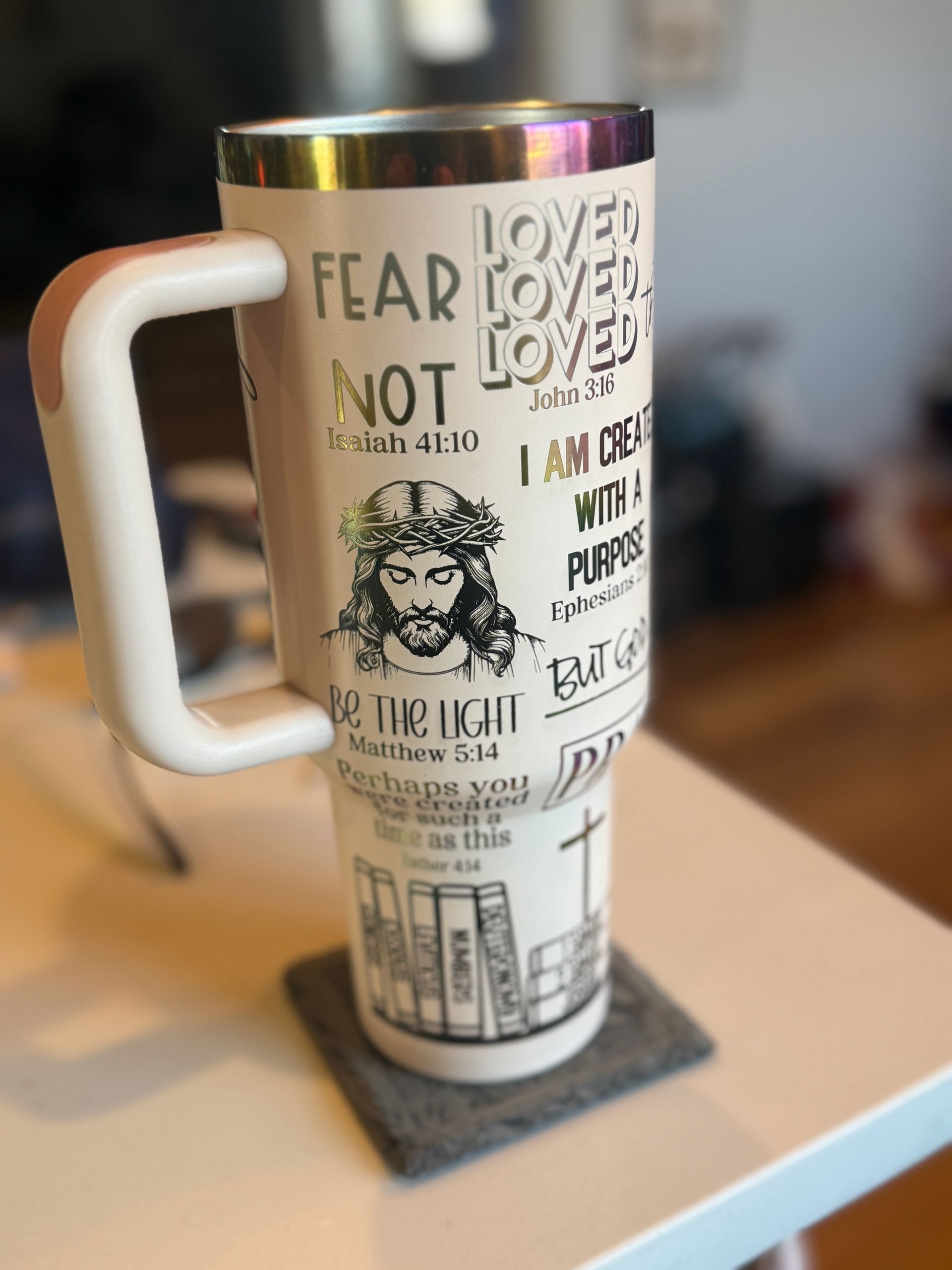 A large white insulated mug with a handle featuring religious quotes and an illustration of Jesus, sitting on a gray coaster.