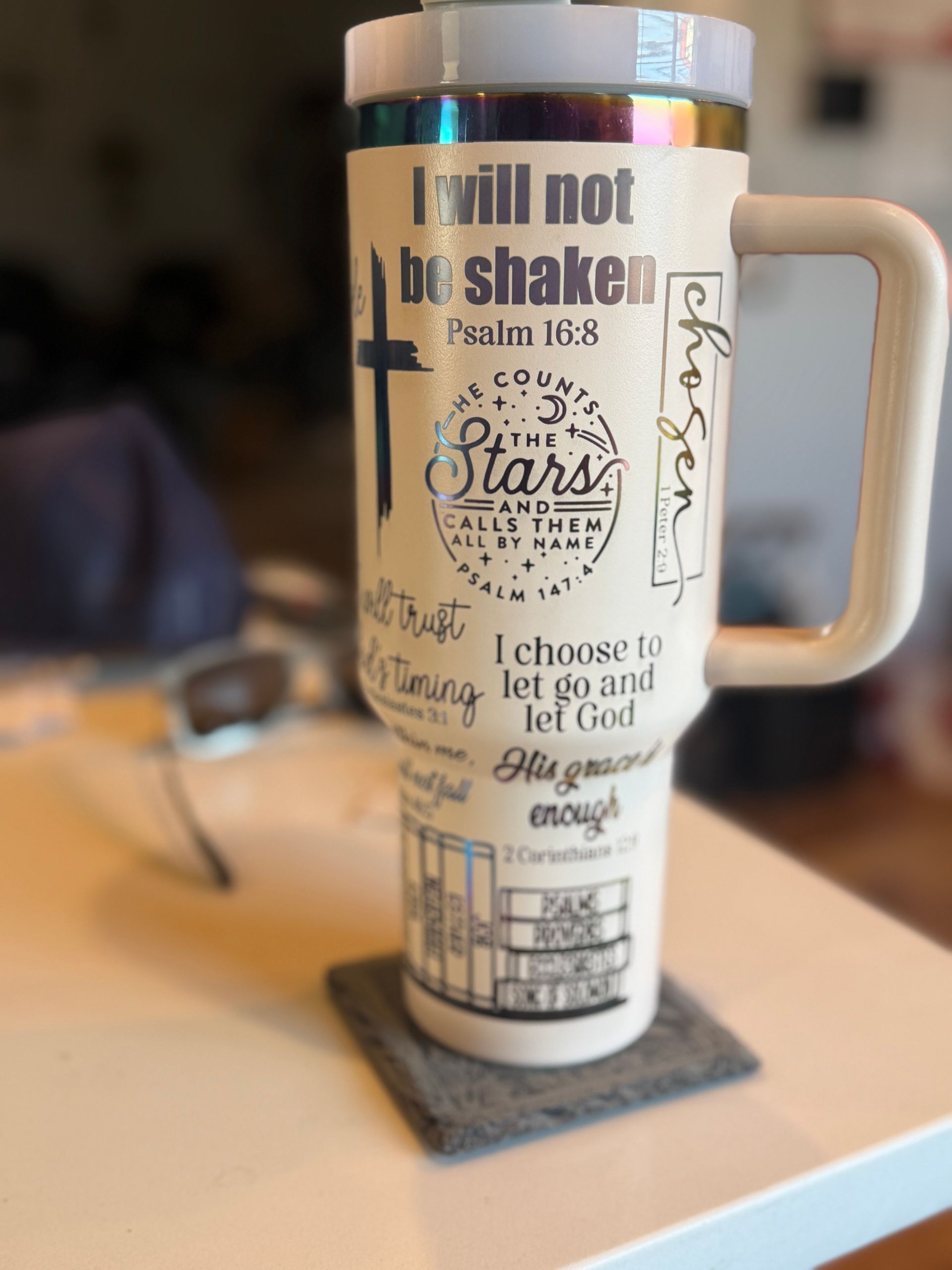 A beige insulated tumbler with inspirational quotes, a cross, and a handle, sitting on a gray coaster.