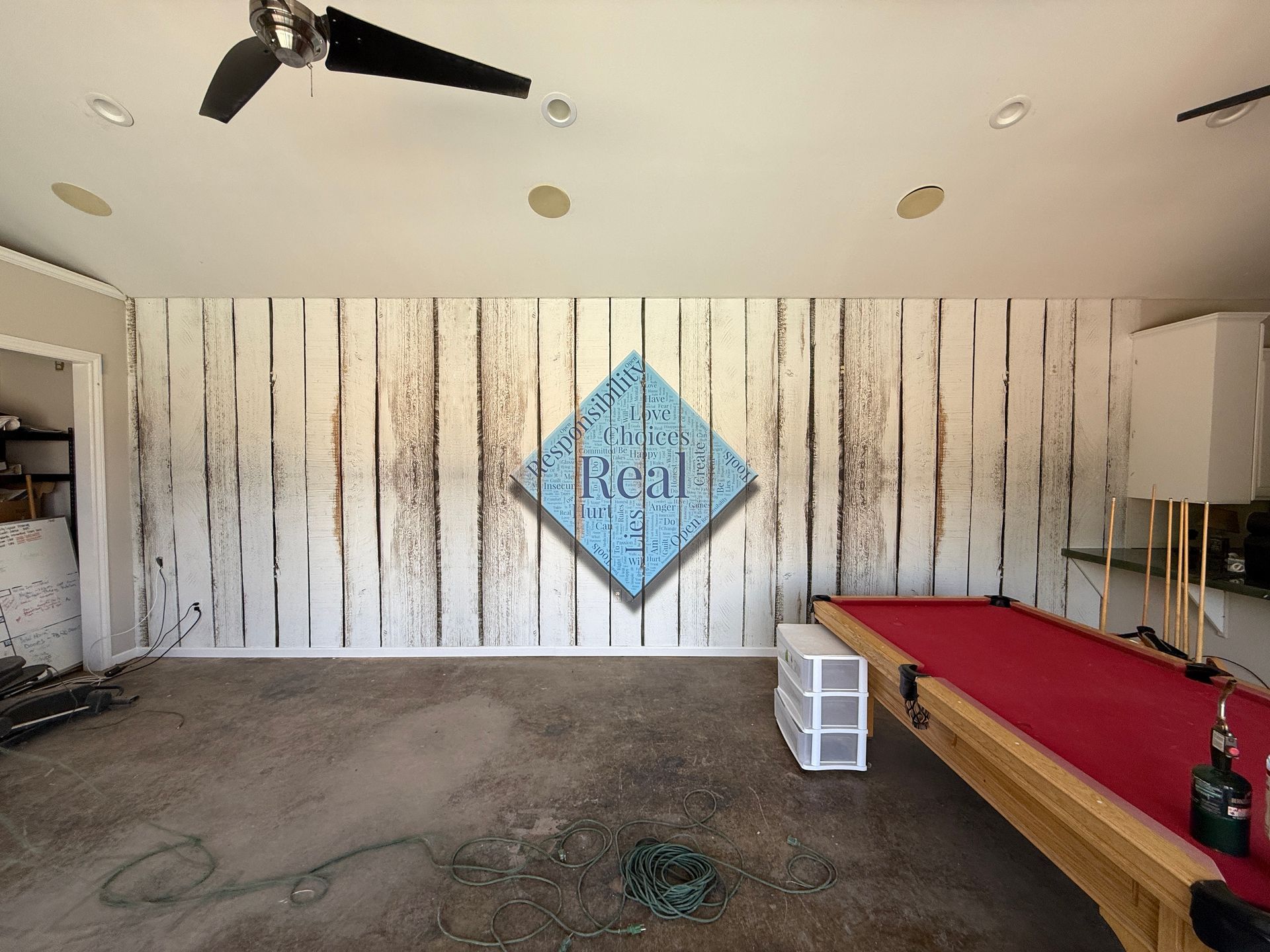 A room under construction features a pool table, a white plank wall, and ceiling fan. A logo is overlaid on the wall.