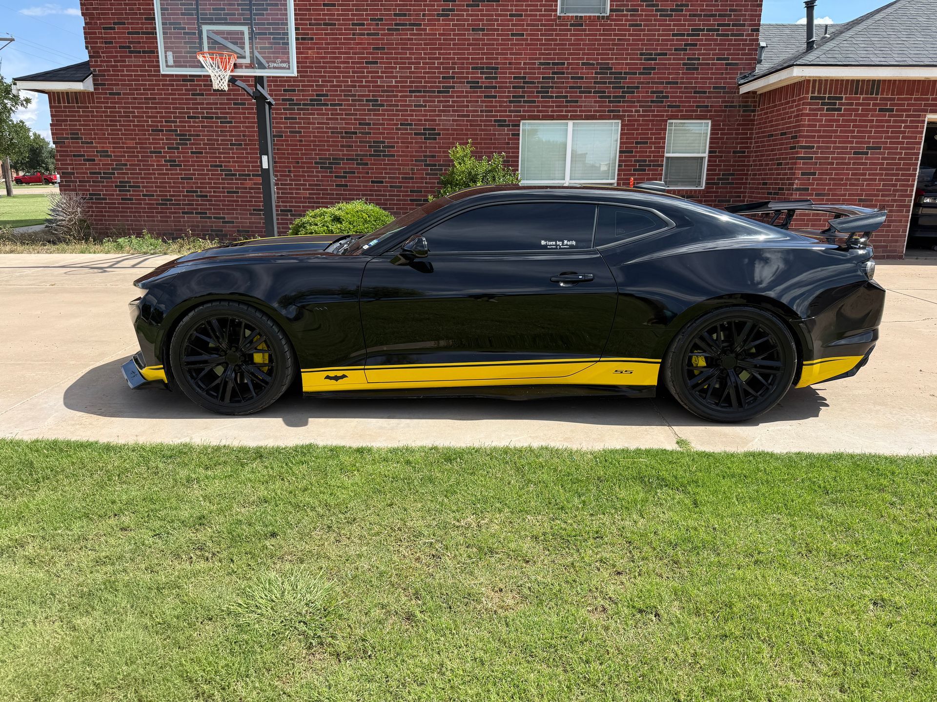 Black sports car with yellow side stripes parked on grass in front of a brick building.