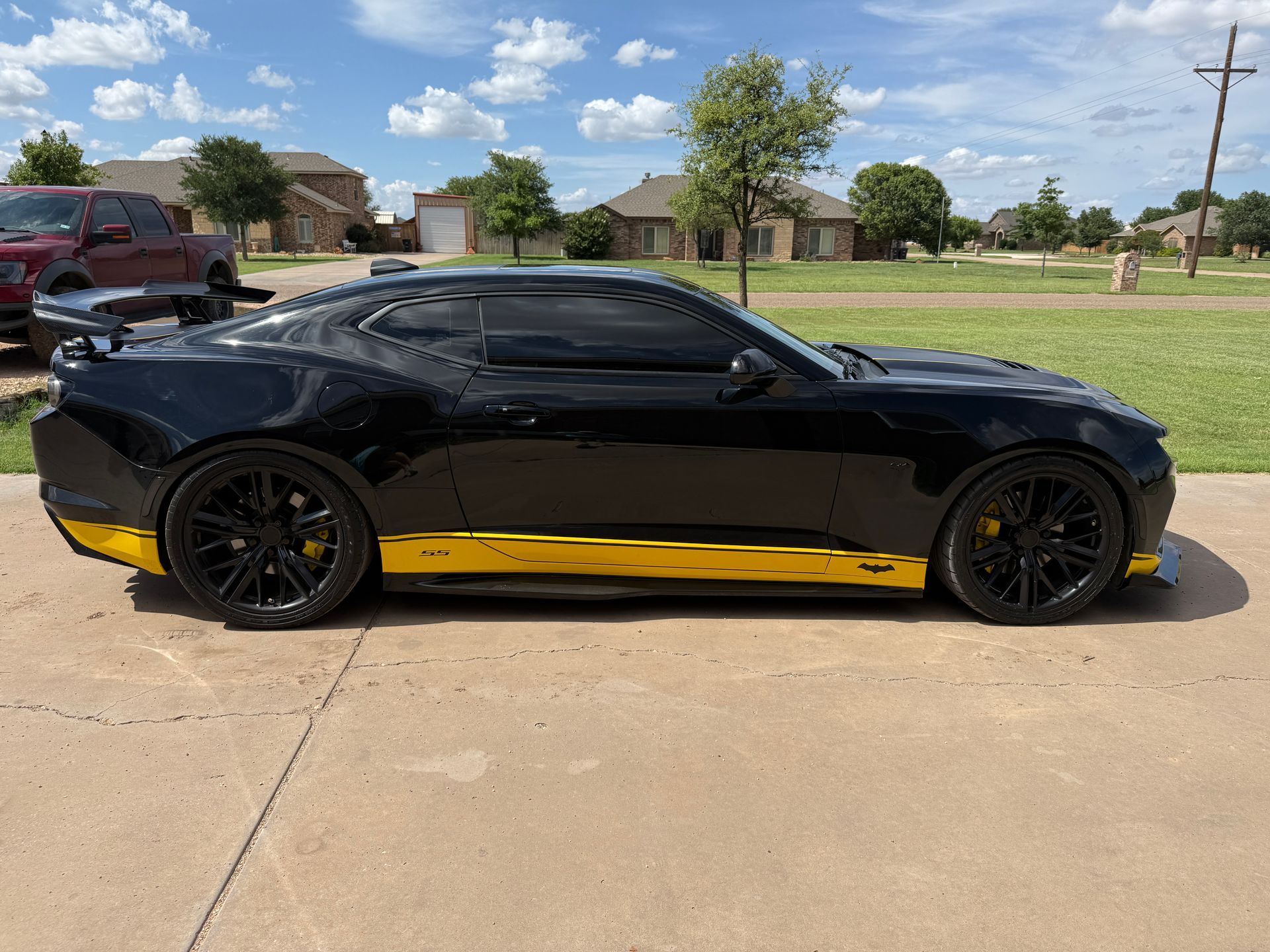 Black sports car with yellow accents parked on a driveway, under a sunny sky, with houses in the background.