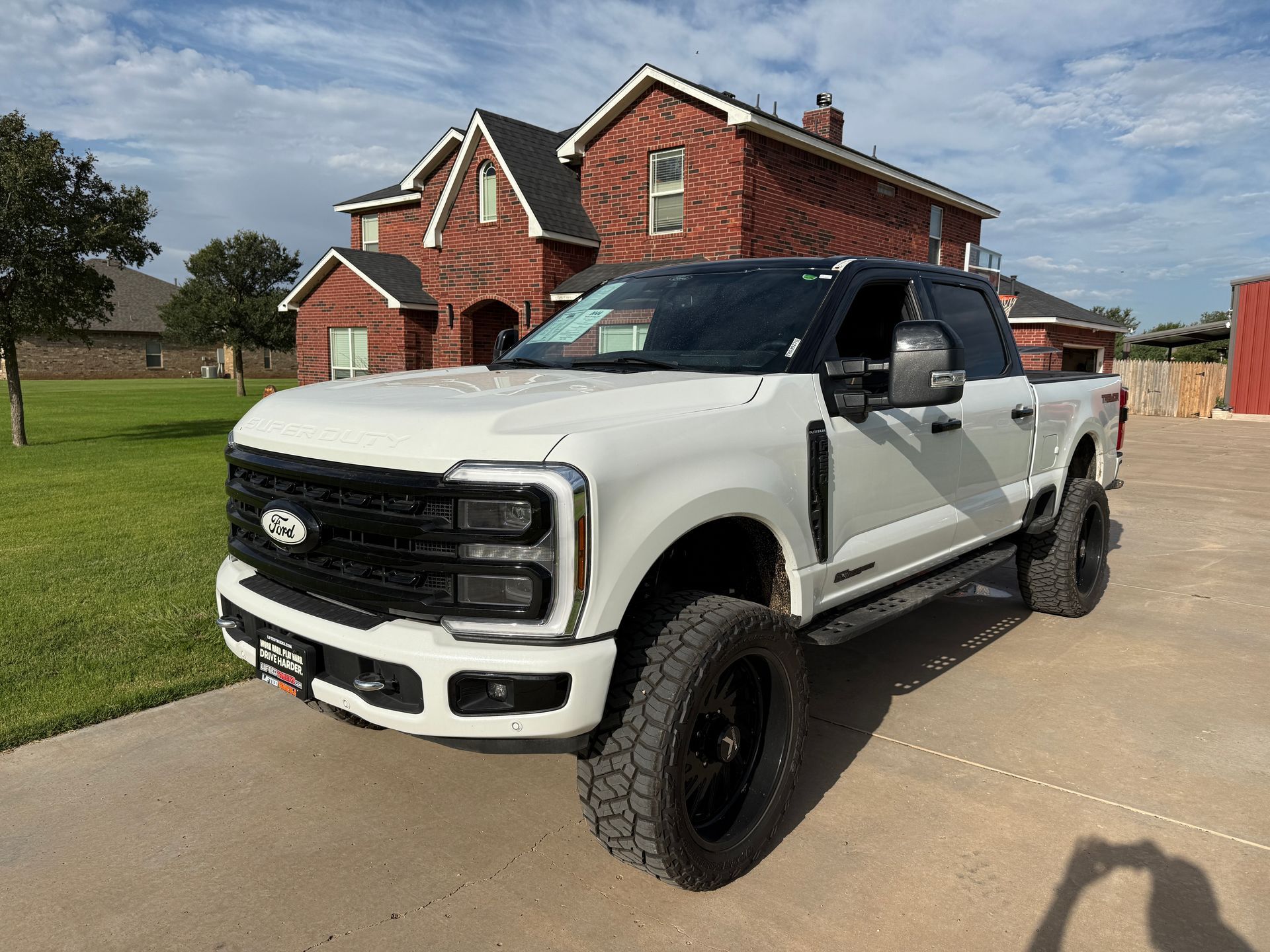 White Ford pickup truck parked in front of a brick house on a sunny day. It has black wheels and a black roof.