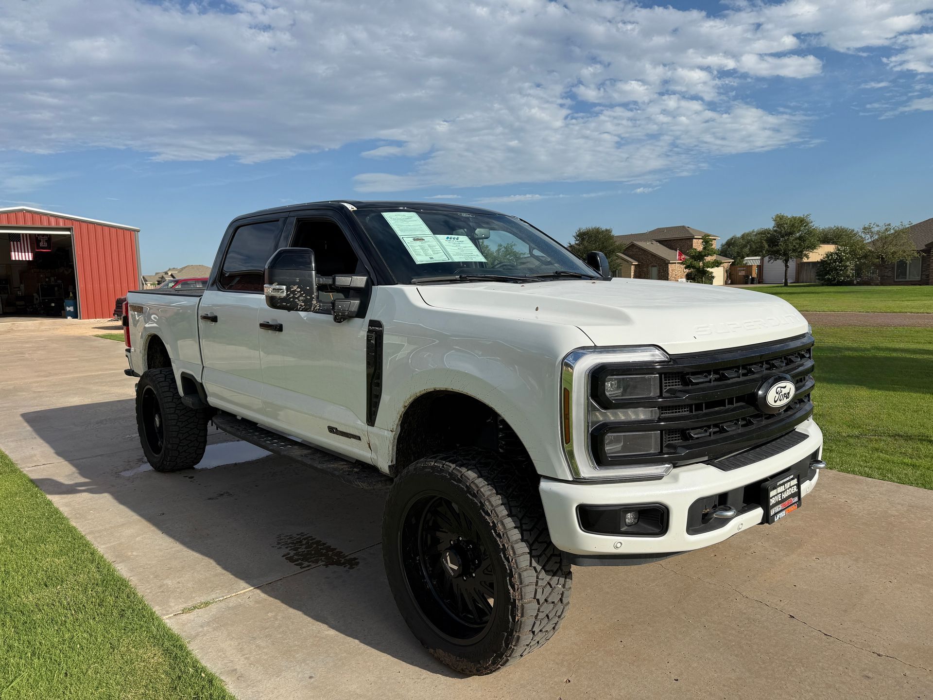 White Ford truck with black accents and large black tires parked on a driveway, with a red building in the background.