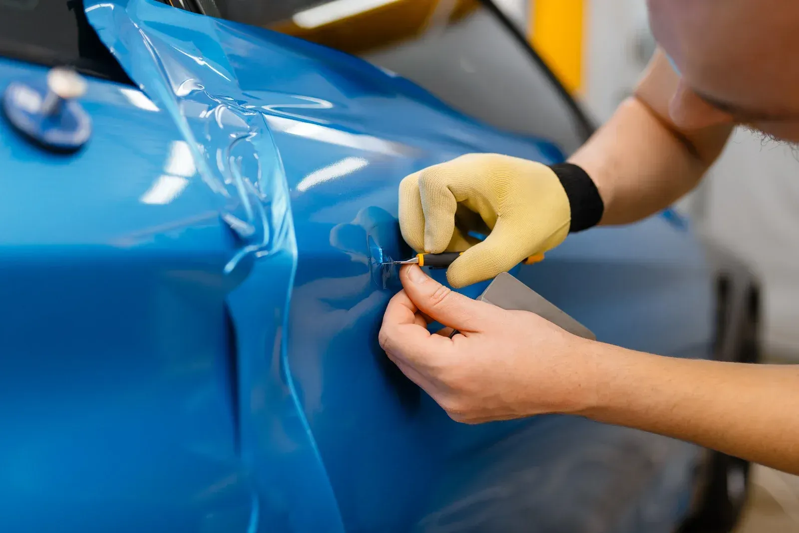 Person in gloves applying blue vinyl wrap to a car door with a small knife.