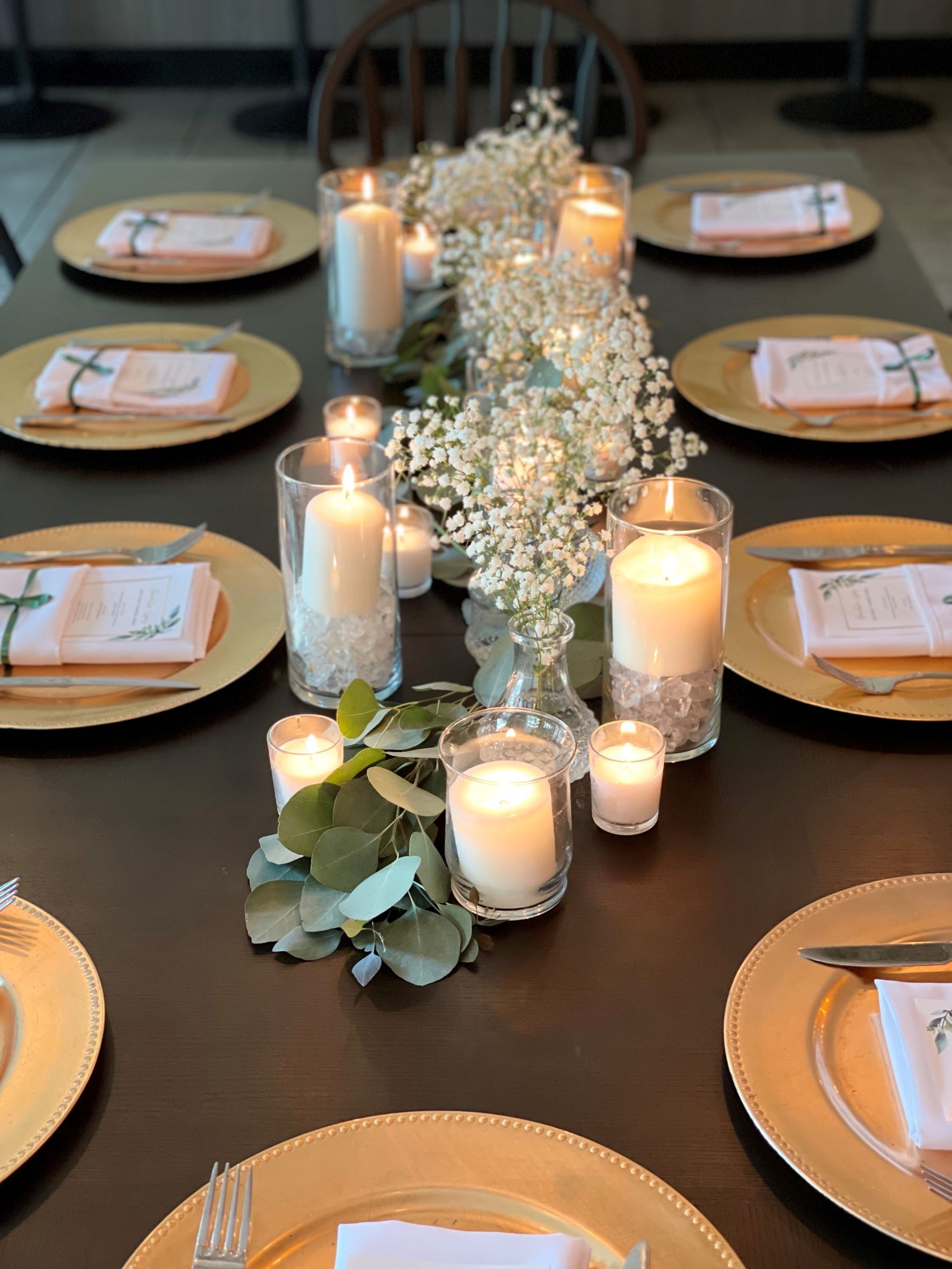 Candles in glass holders next to white flowers in small vases on rehearsal dinner table with eucalyptus branches, gold plates and silver utensils at The Nook in Temecula, CA