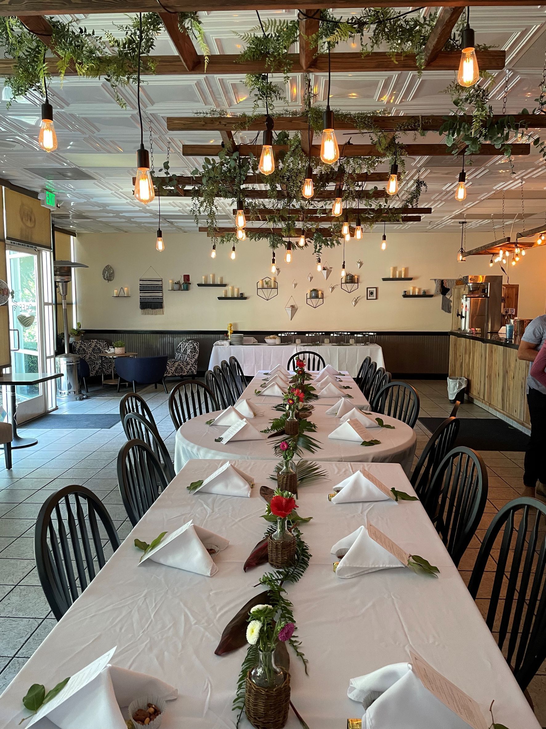 A long table covered in white linens with folded napkins and red flowers surrounded by green foliage decorate the table for a rehearsal dinner at The Nook in Temecula, CA hosted by Wine Country Catering