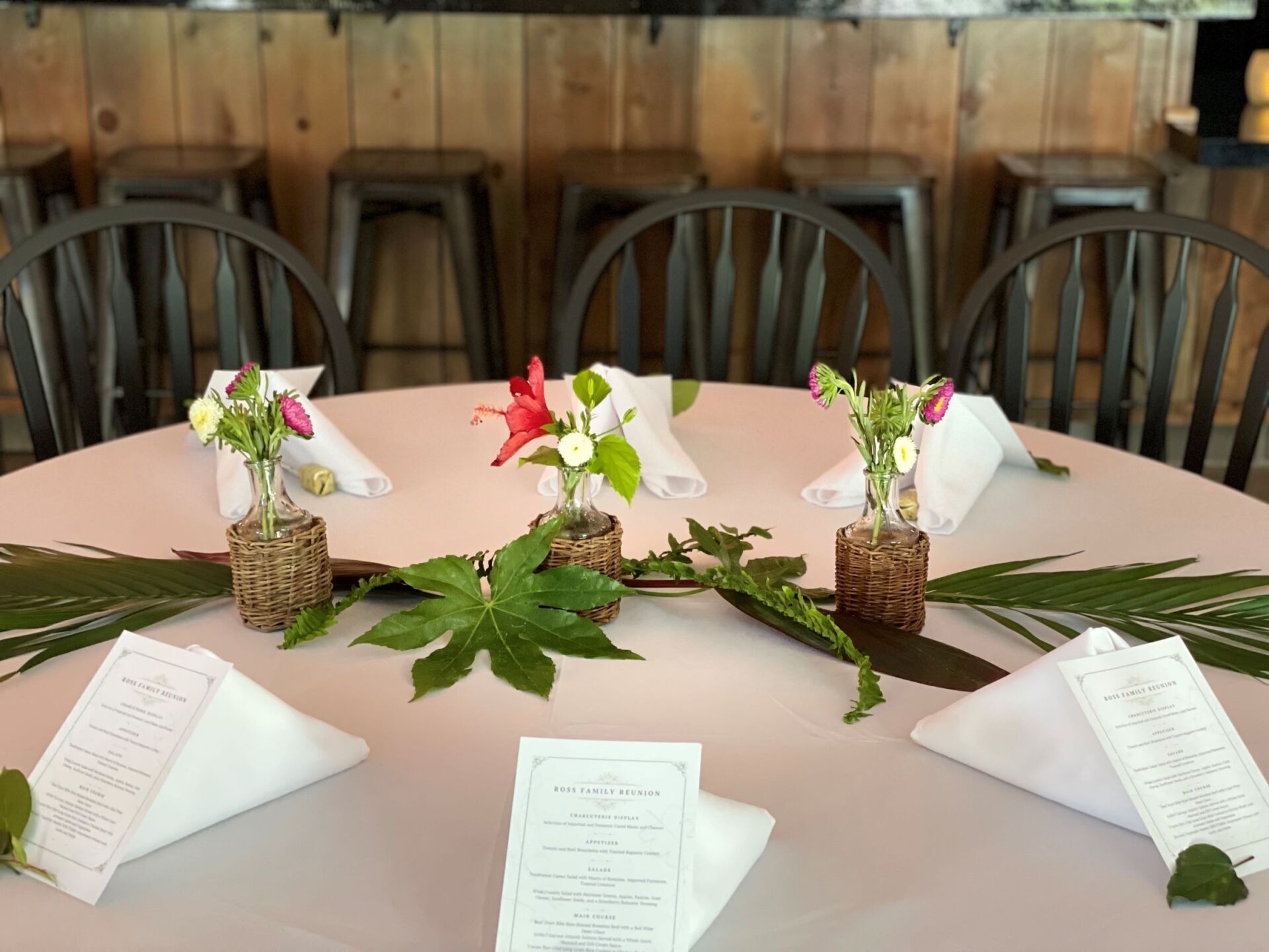 Cut flowers in wicker wrapped vases with folded white napkins and palm fronds decorate table for a rehearsal dinner at The Nook in Temecula, CA