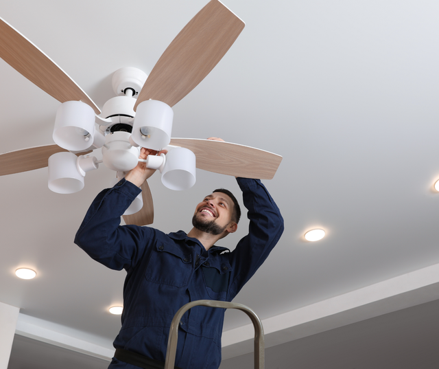 Man in blue jumpsuit installing a ceiling fan on a ladder, smiling. White ceiling, light-colored blades.