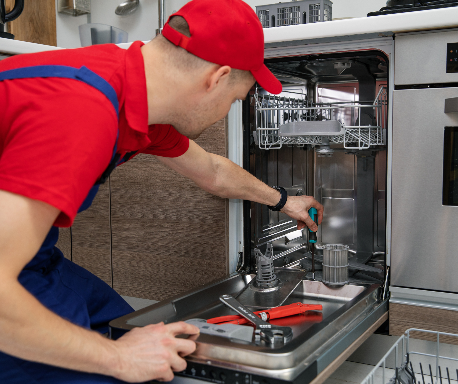 Man in red cap and shirt repairing a dishwasher; kitchen setting.