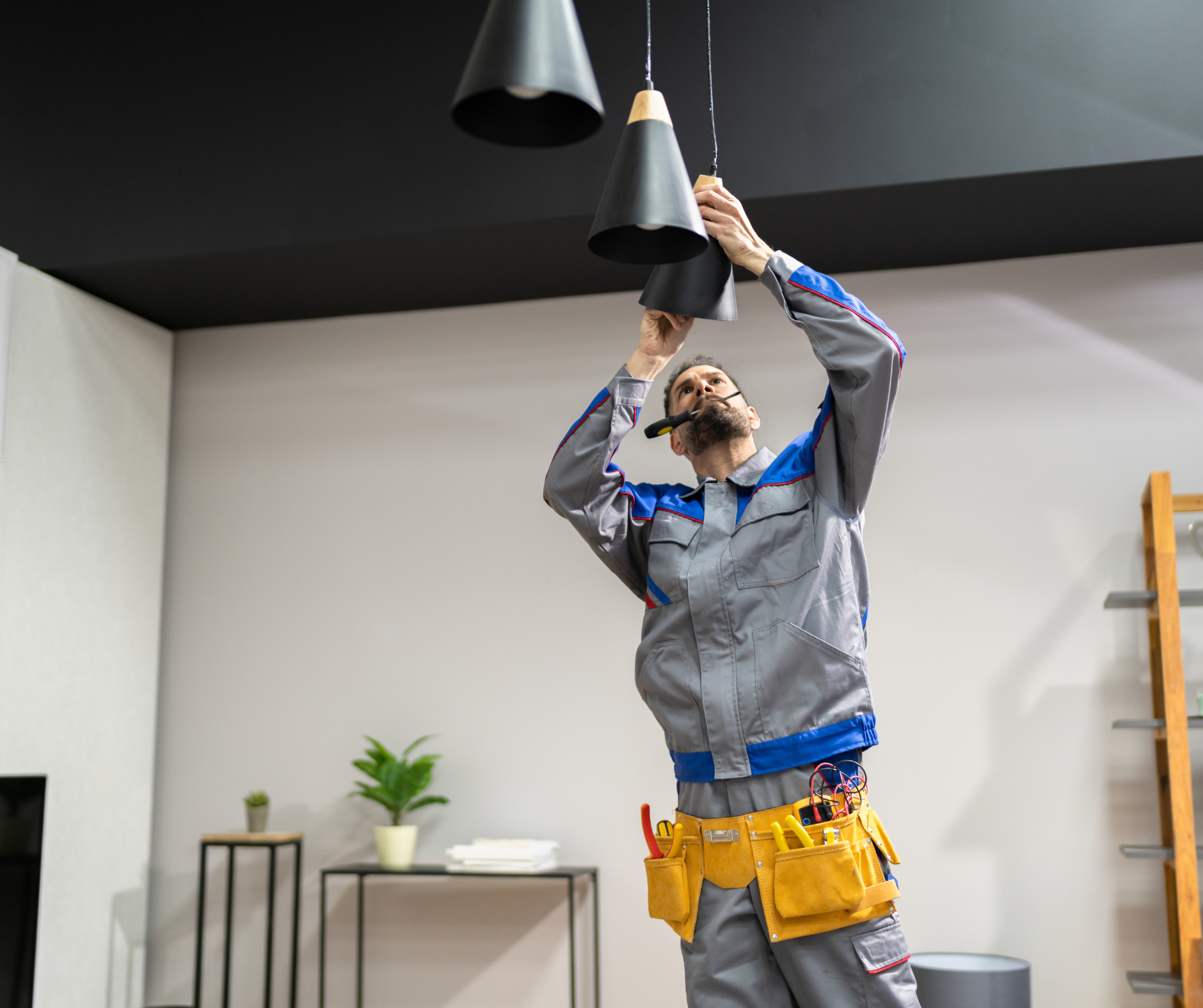 Electrician installing a pendant light fixture in a room with a black ceiling; they're wearing work clothes, tools visible.