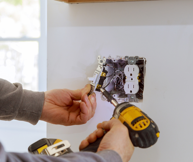 Person installing electrical outlet with a drill; white wall, visible wires.