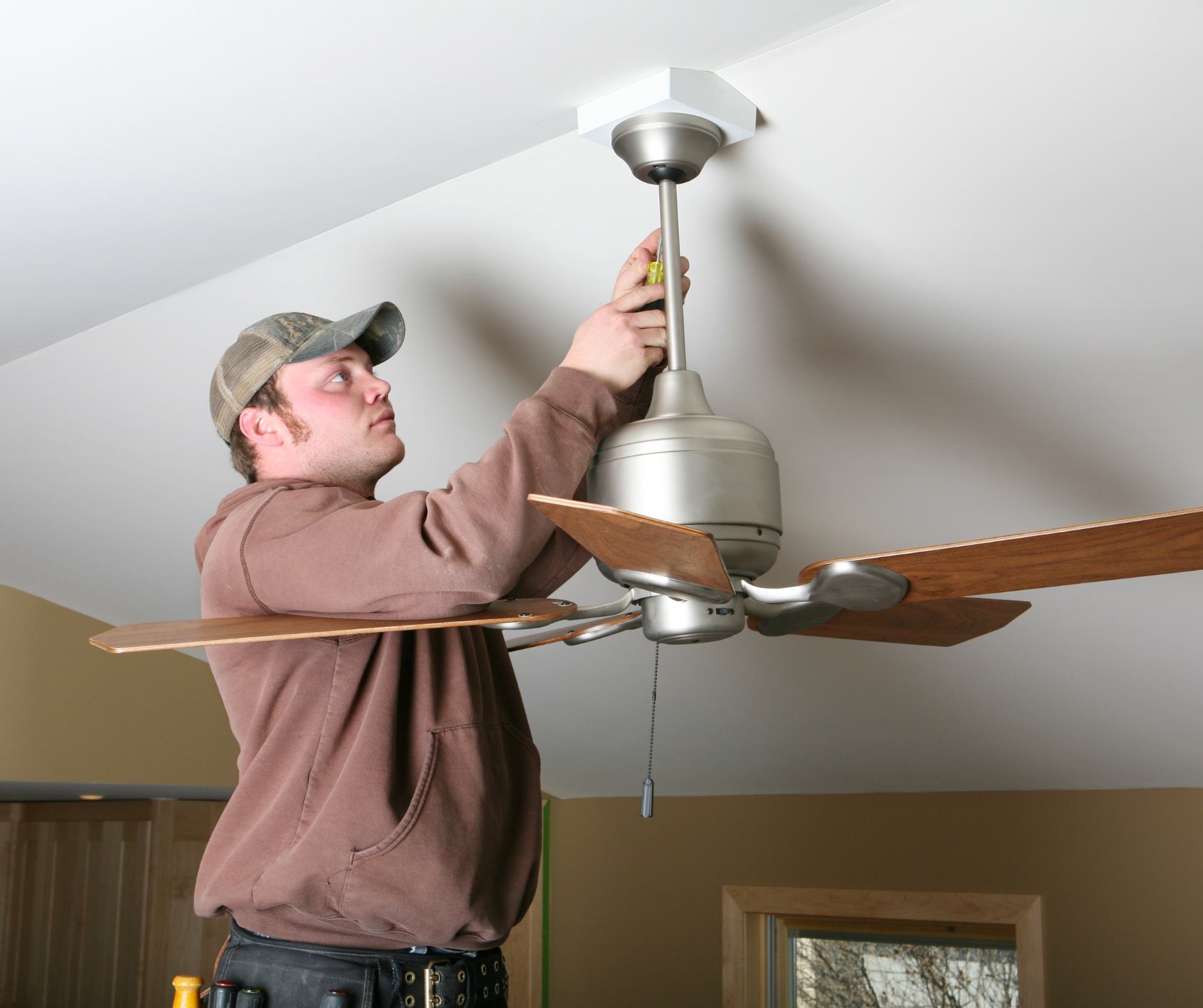Man in cap installing a ceiling fan. He's on a ladder, working in a room with a white ceiling.