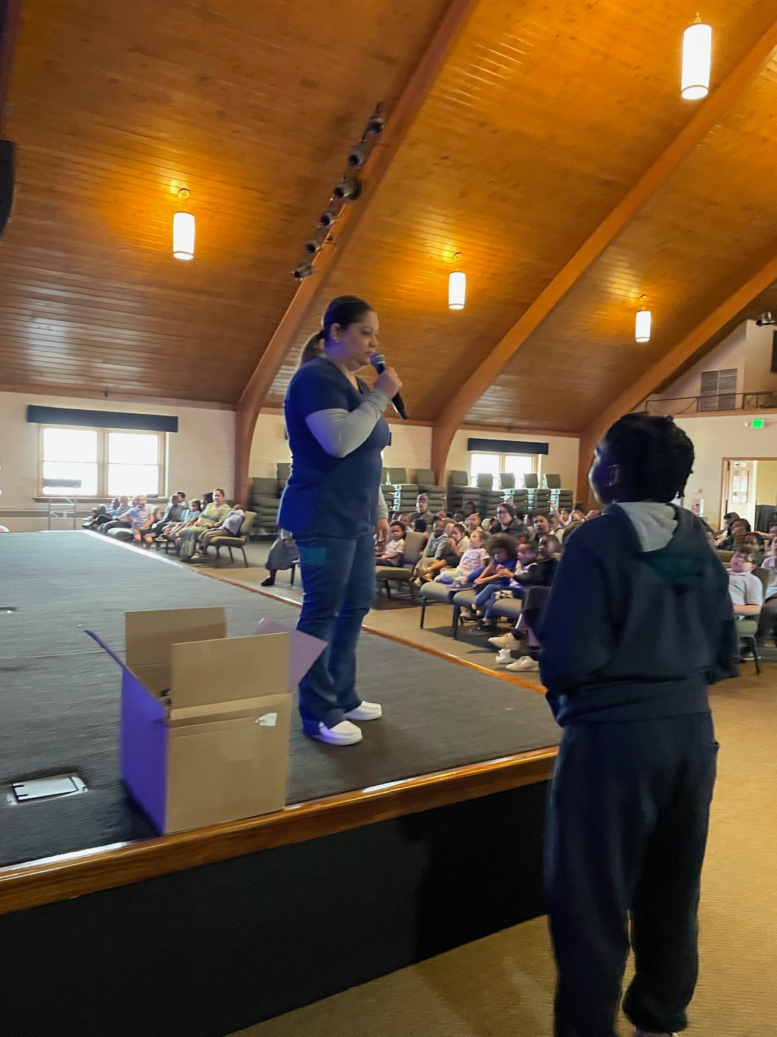 A woman is standing on a stage talking into a microphone while a boy stands behind her.